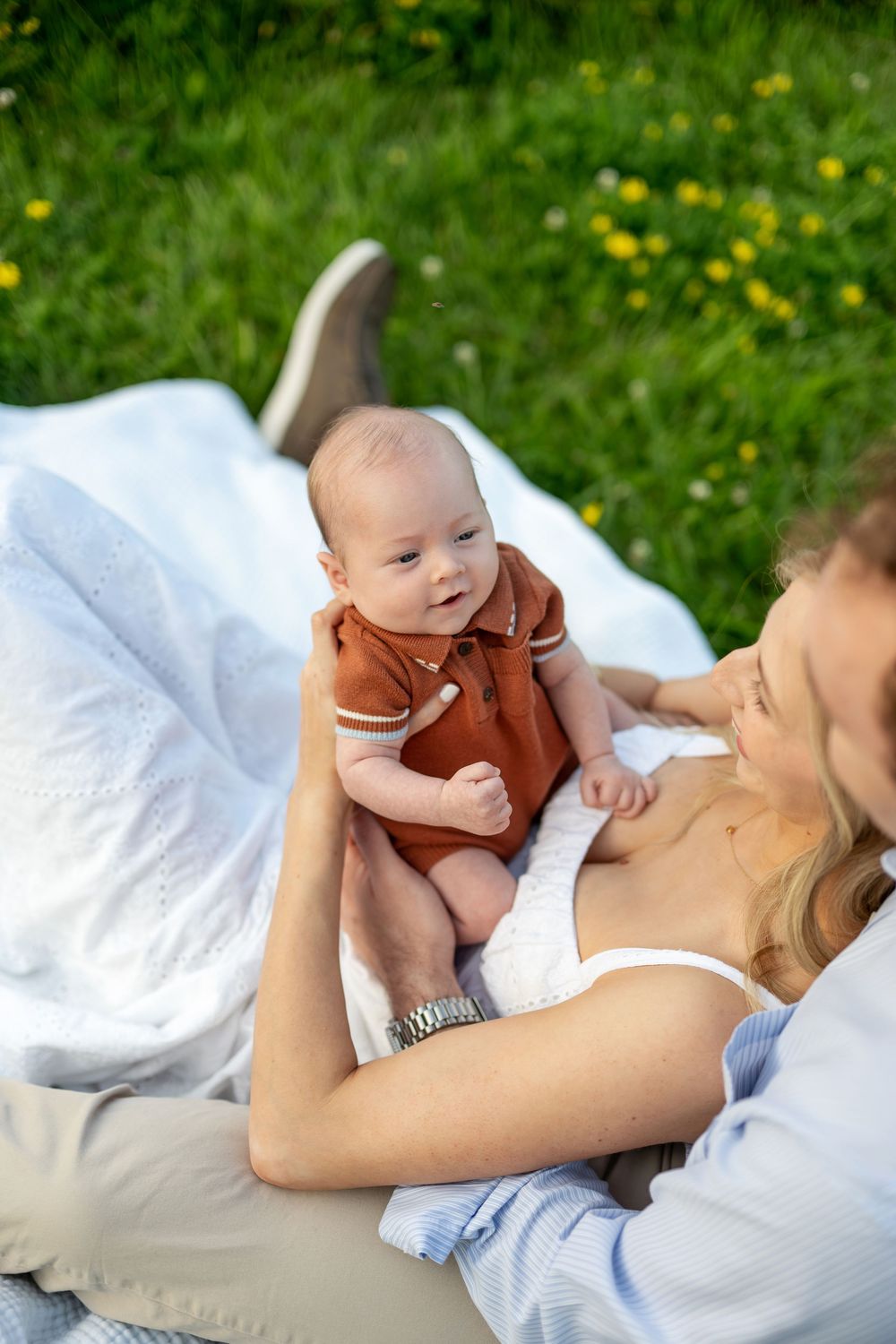 Mother lying on blanket in grass, lovingly holding her infant baby dressed in a rust-colored romper outdoors.