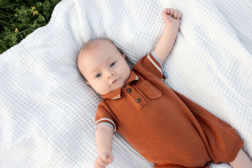 A cute baby in a rust-colored polo romper lies on a white blanket outdoors, reaching up with one arm.