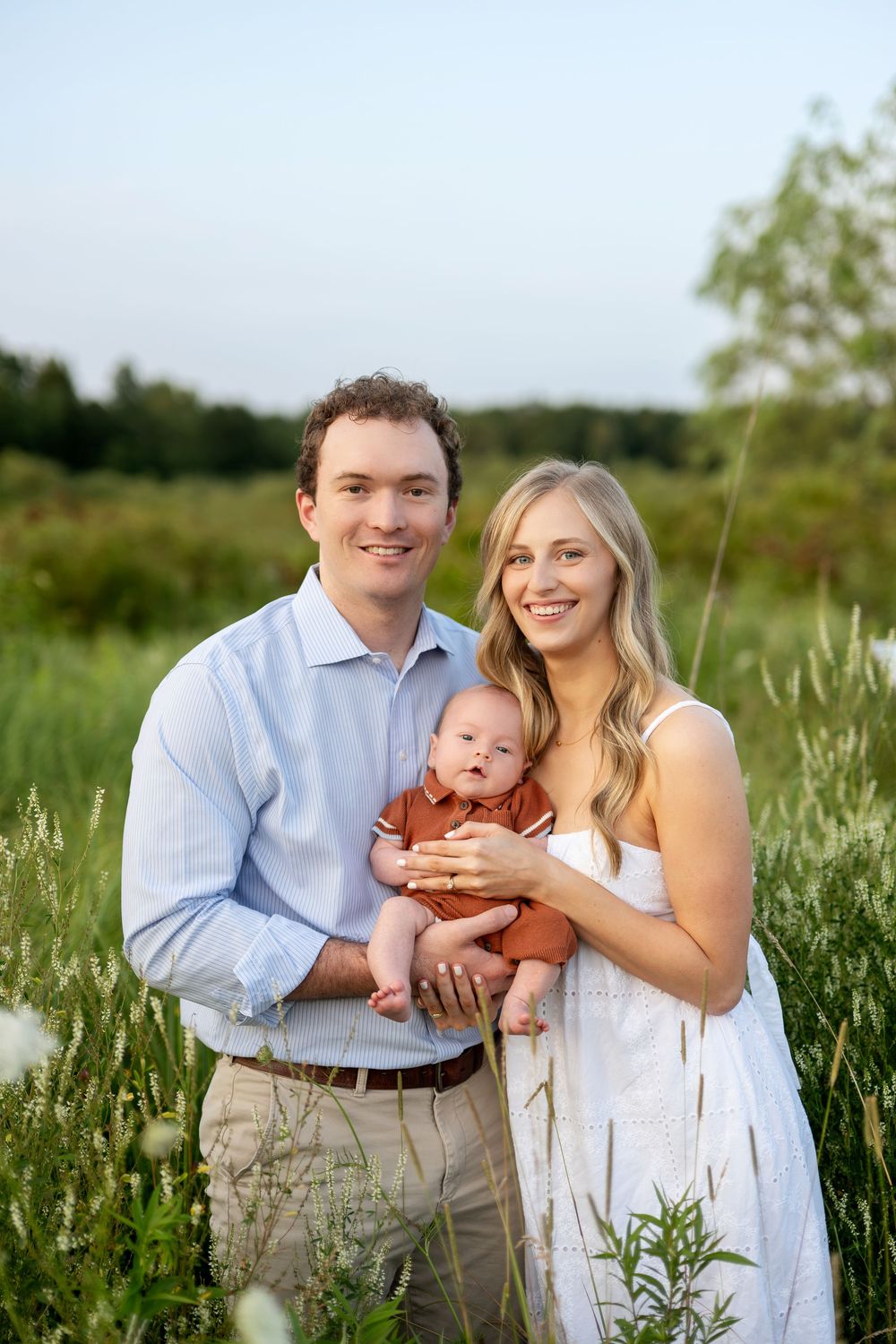Smiling young family with newborn baby posing together in a beautiful green field at sunset.