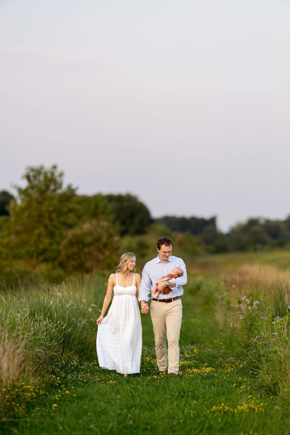 A couple walks hand-in-hand through a green meadow, the man cradling a newborn baby, during a family outdoor photo session.