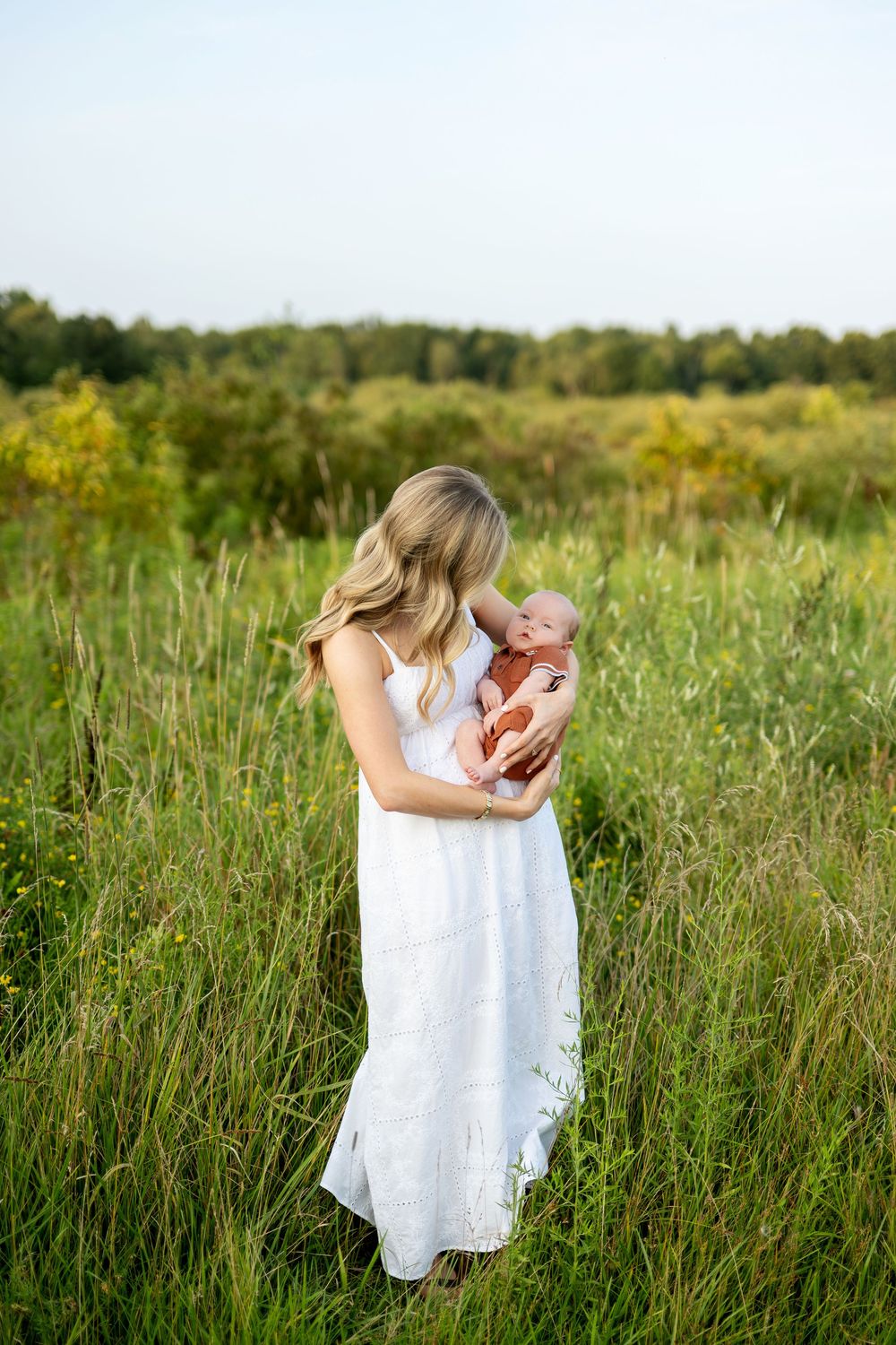 A blonde woman in a white dress holds her newborn baby in a lush green meadow field.