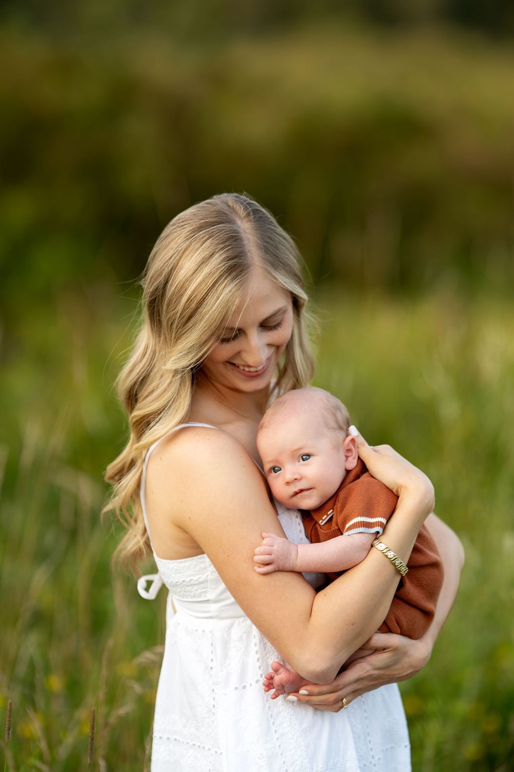 Smiling blonde mother in white dress tenderly holding newborn baby in rust romper outdoors in green field.