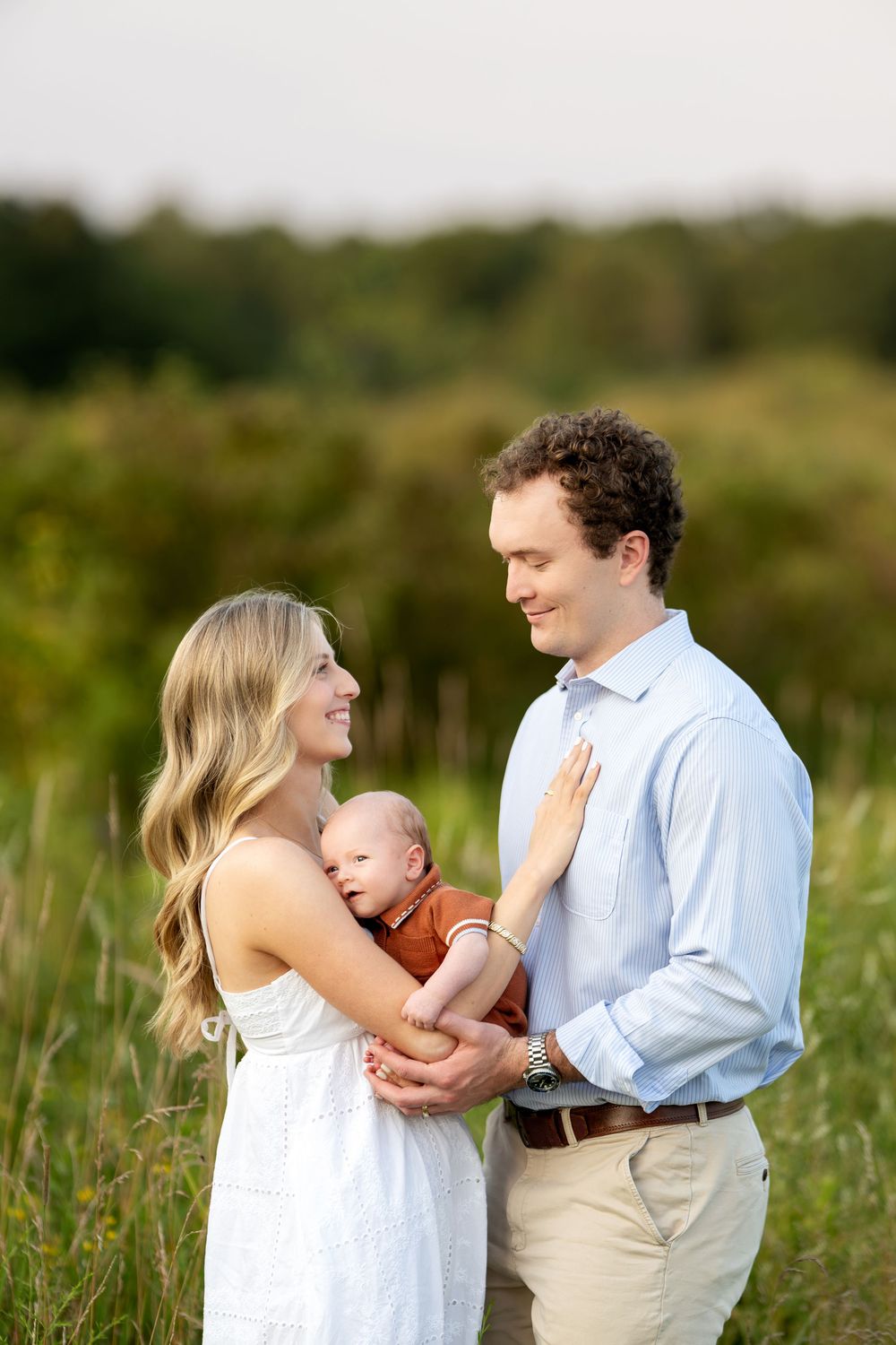 Young couple smiling and holding newborn baby in a sunlit green field during a family portrait session.