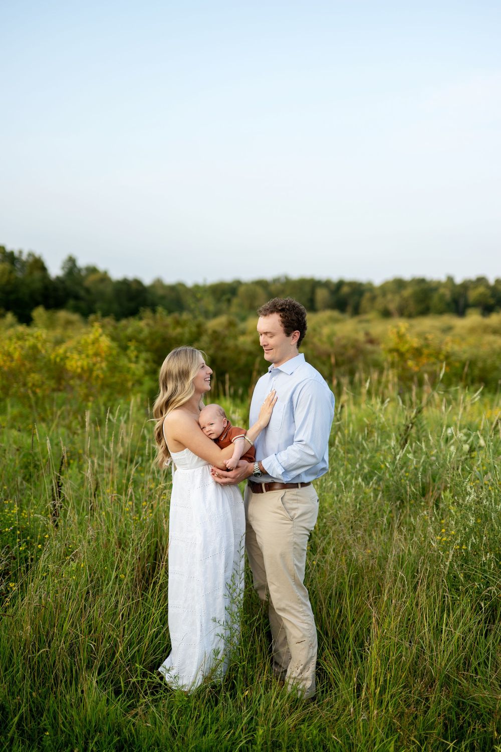 A young family stands in a lush green field, mother in white dress holding newborn as father smiles lovingly.