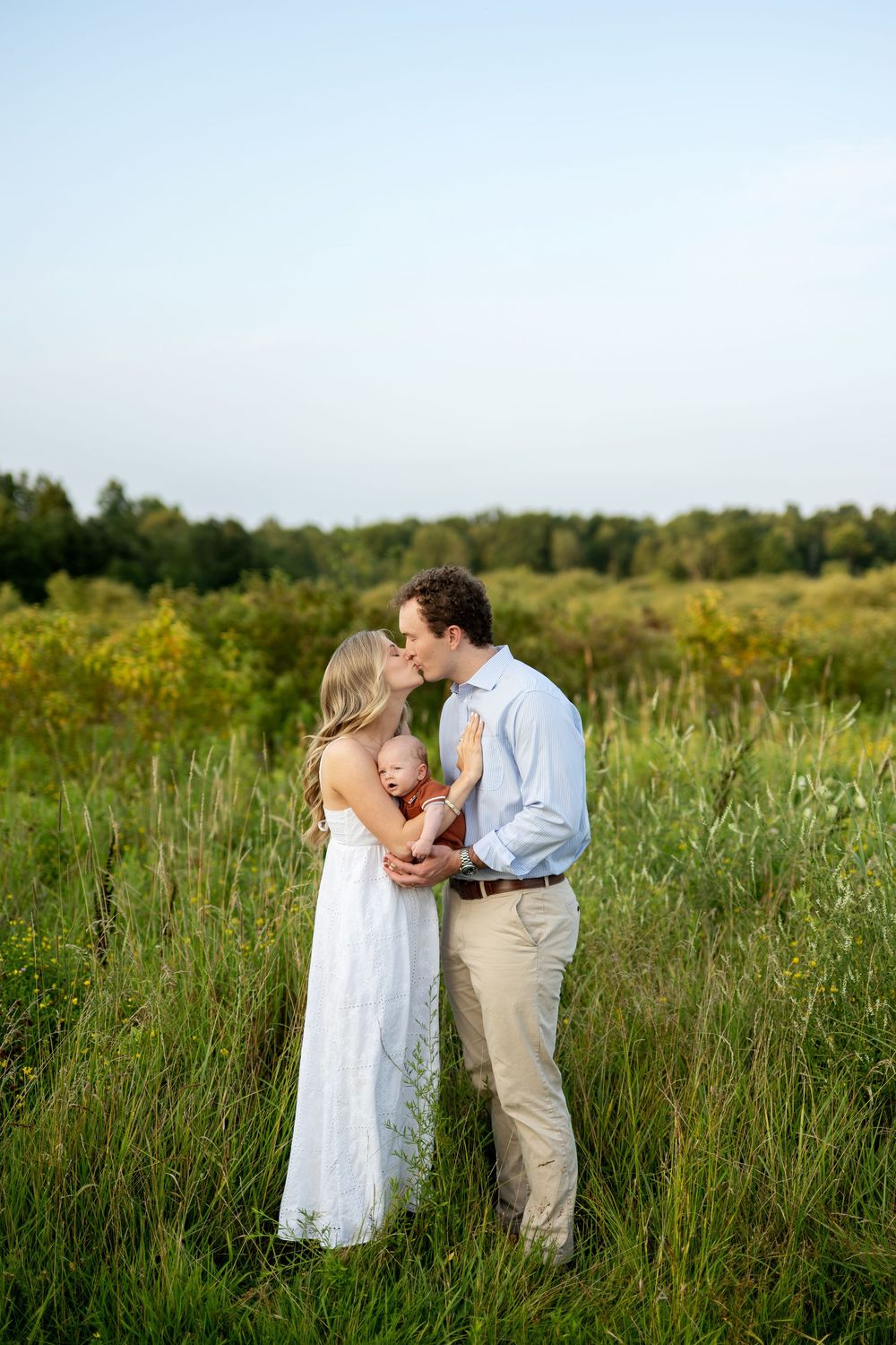 A couple kisses in a green field while holding their newborn baby, the woman in a white dress and man in a blue shirt.