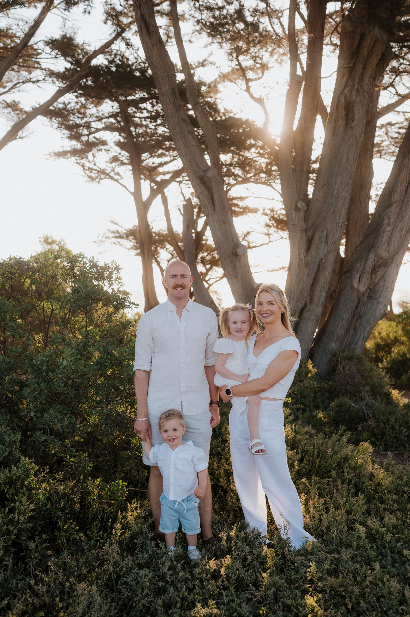 Family of four in white outfits posing outdoors under large trees with golden sunlight in background.