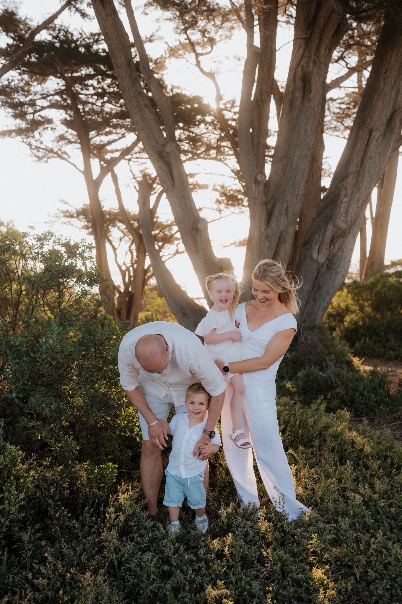 Family of four posing outdoors near a large tree at golden hour, parents and two young children dressed in white.