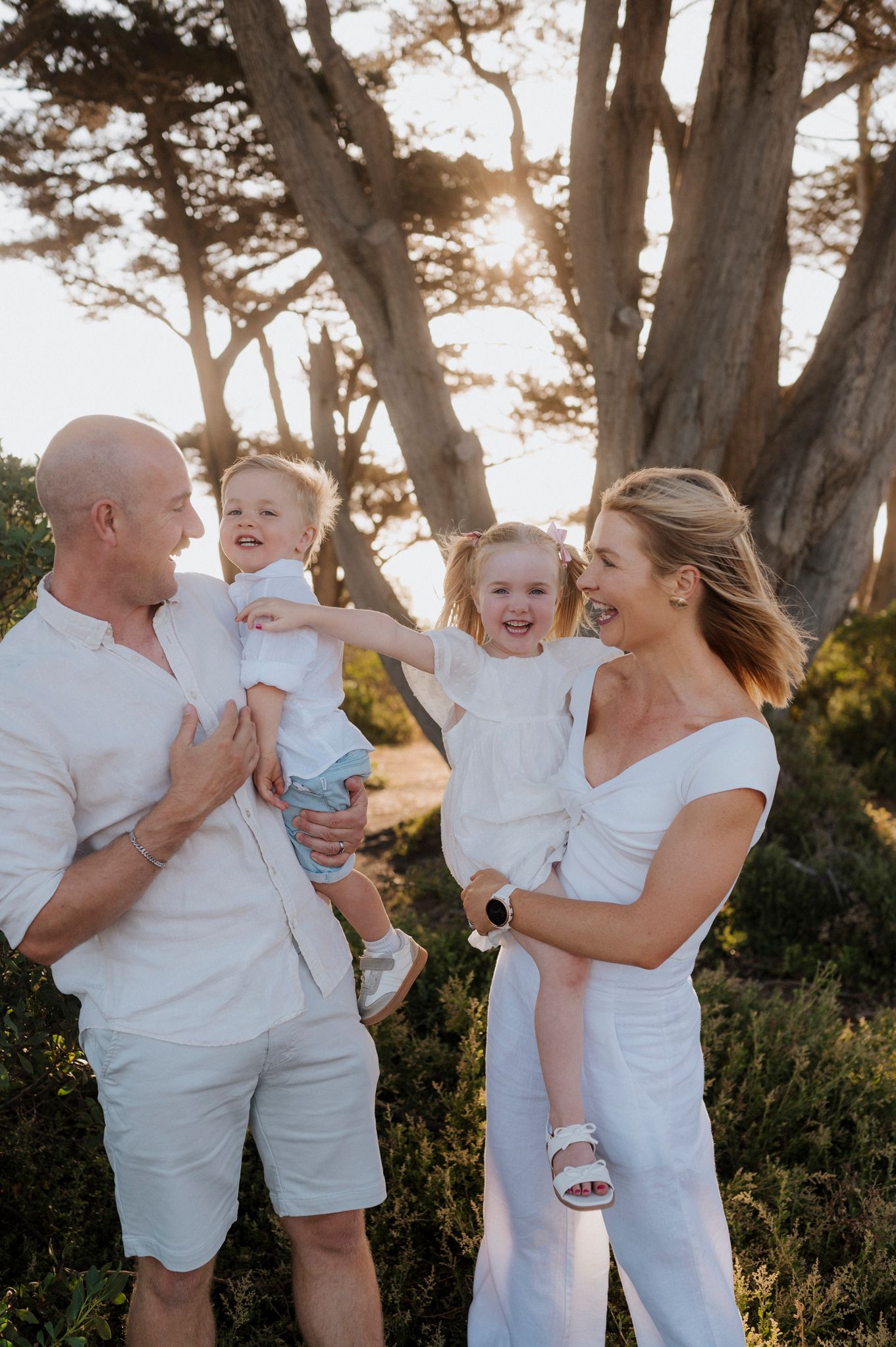 Happy family of four in white outfits laughing outdoors with sunlit trees in background.