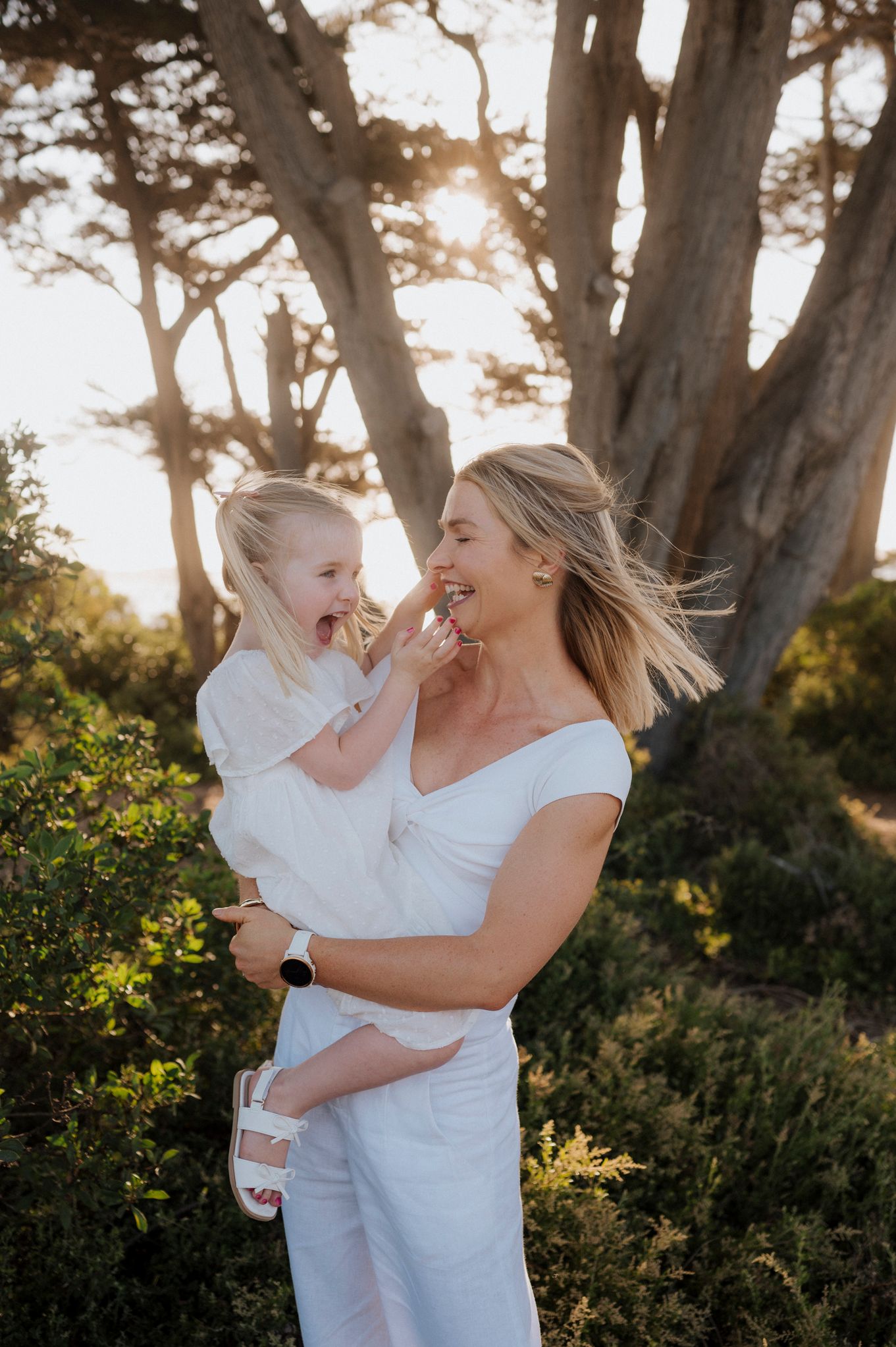 A mother in white holds her laughing young daughter outdoors, both in white outfits, with sunlit trees in the background.