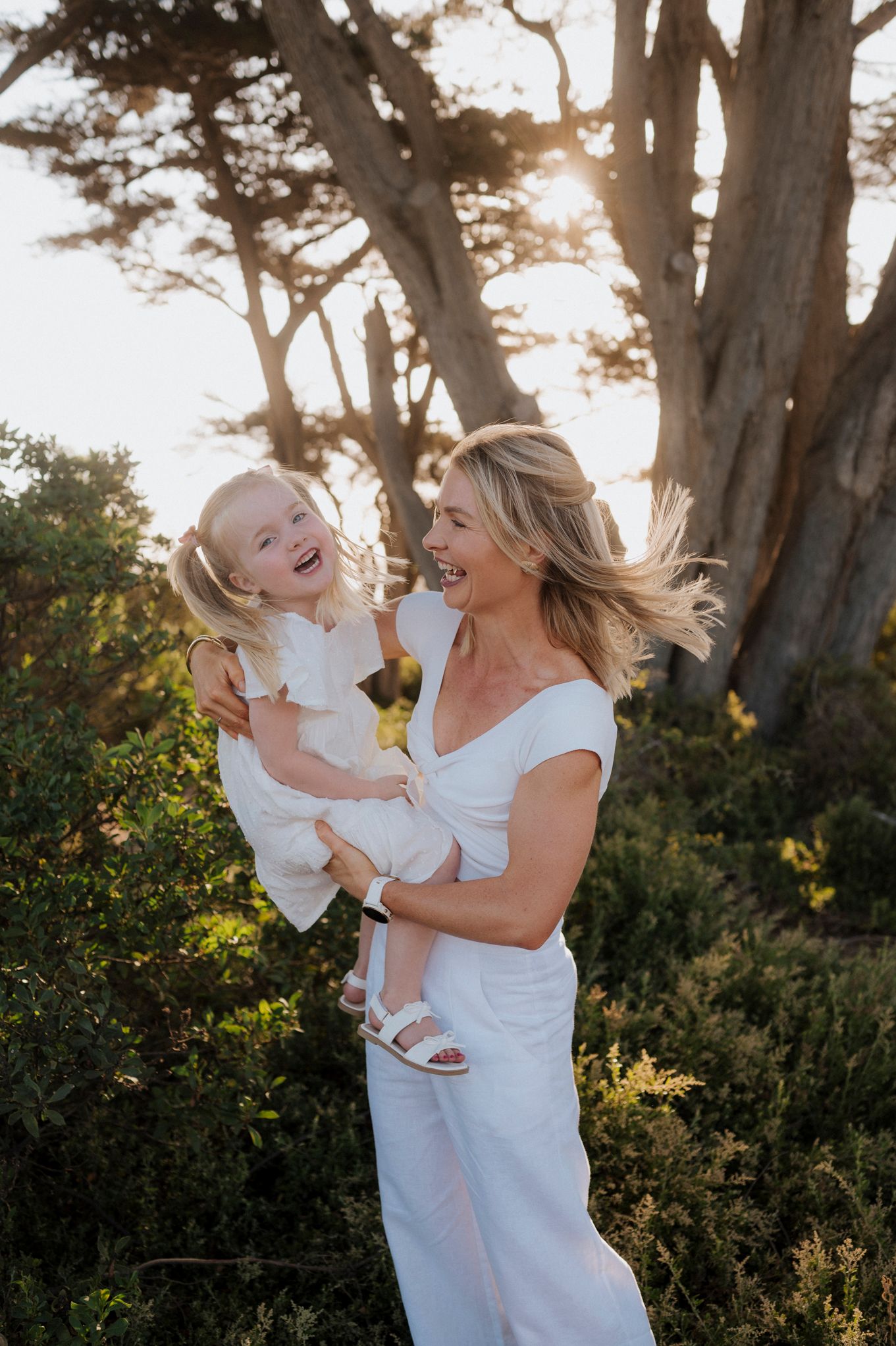 A joyful woman in white holds a laughing blonde toddler girl outdoors among green shrubs and trees in golden sunlight.