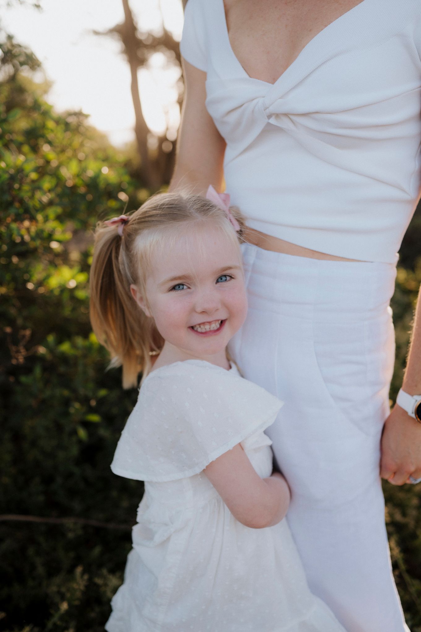 A smiling young girl in a white dress hugs a woman in white outfit outdoors in golden sunlight.
