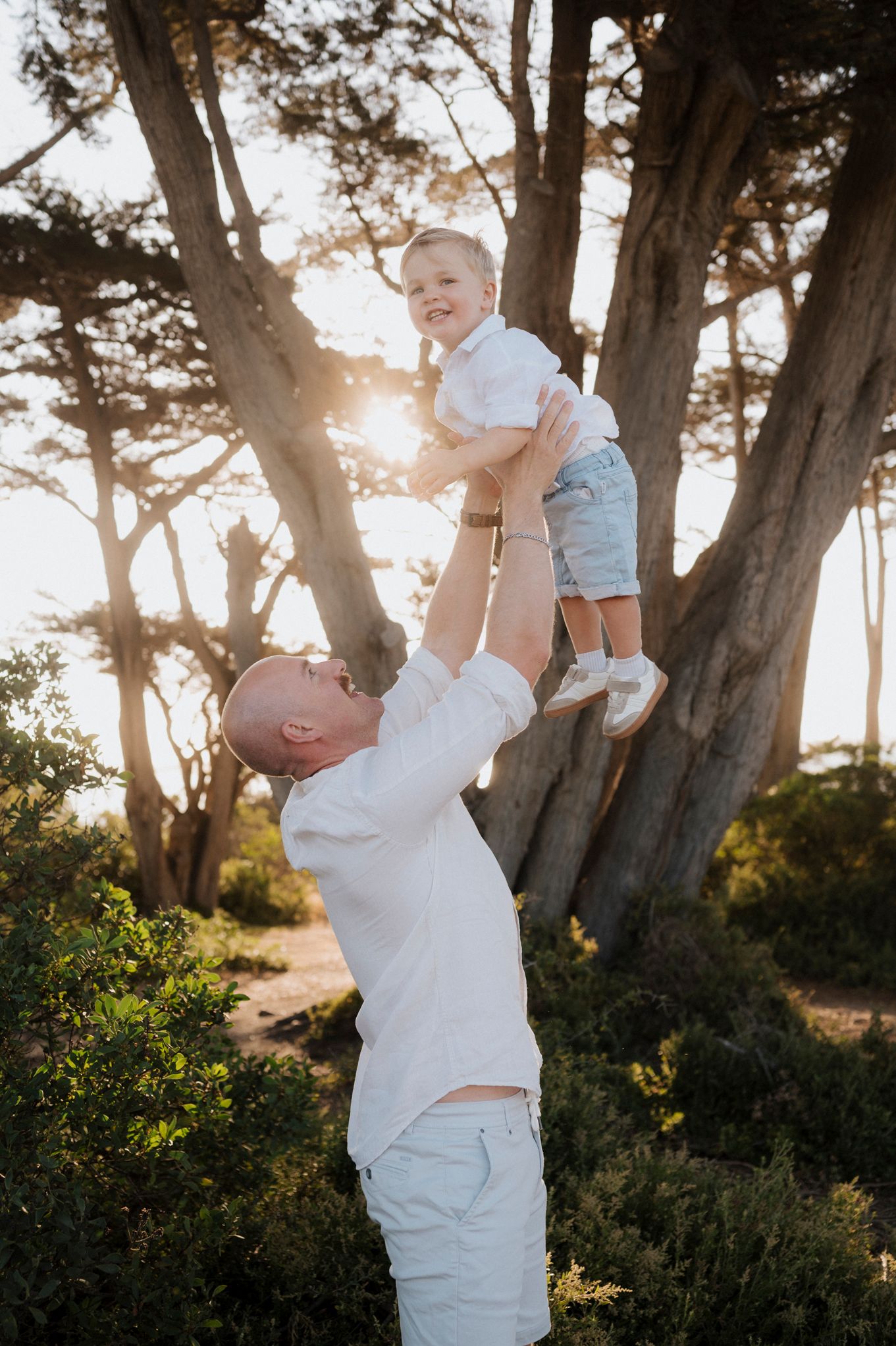 A bald father lifts his smiling toddler son high in the air outdoors, with golden sunlight filtering through tall trees.