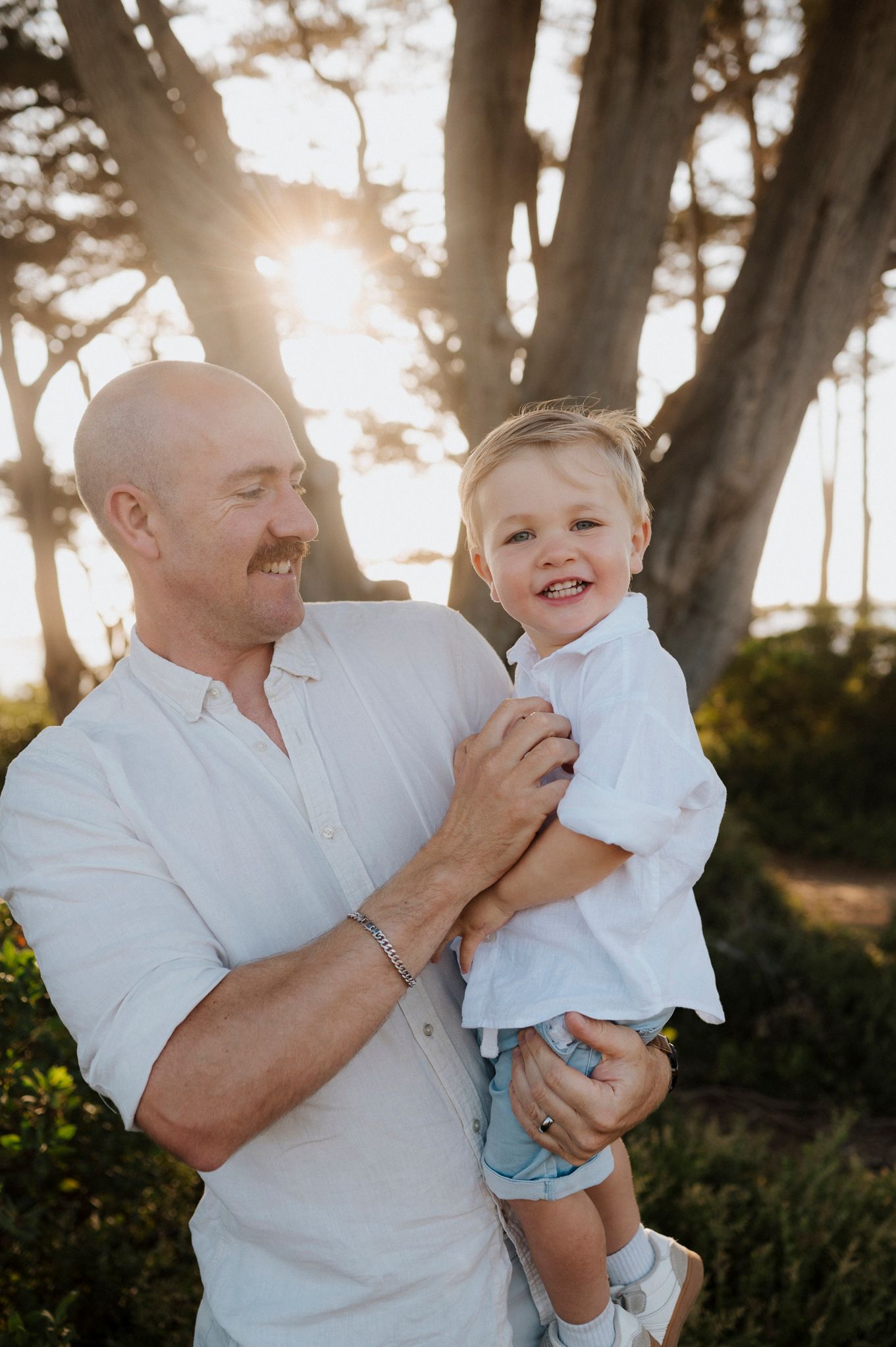 Bald man with mustache holding smiling toddler boy outdoors among trees with golden sunlight.