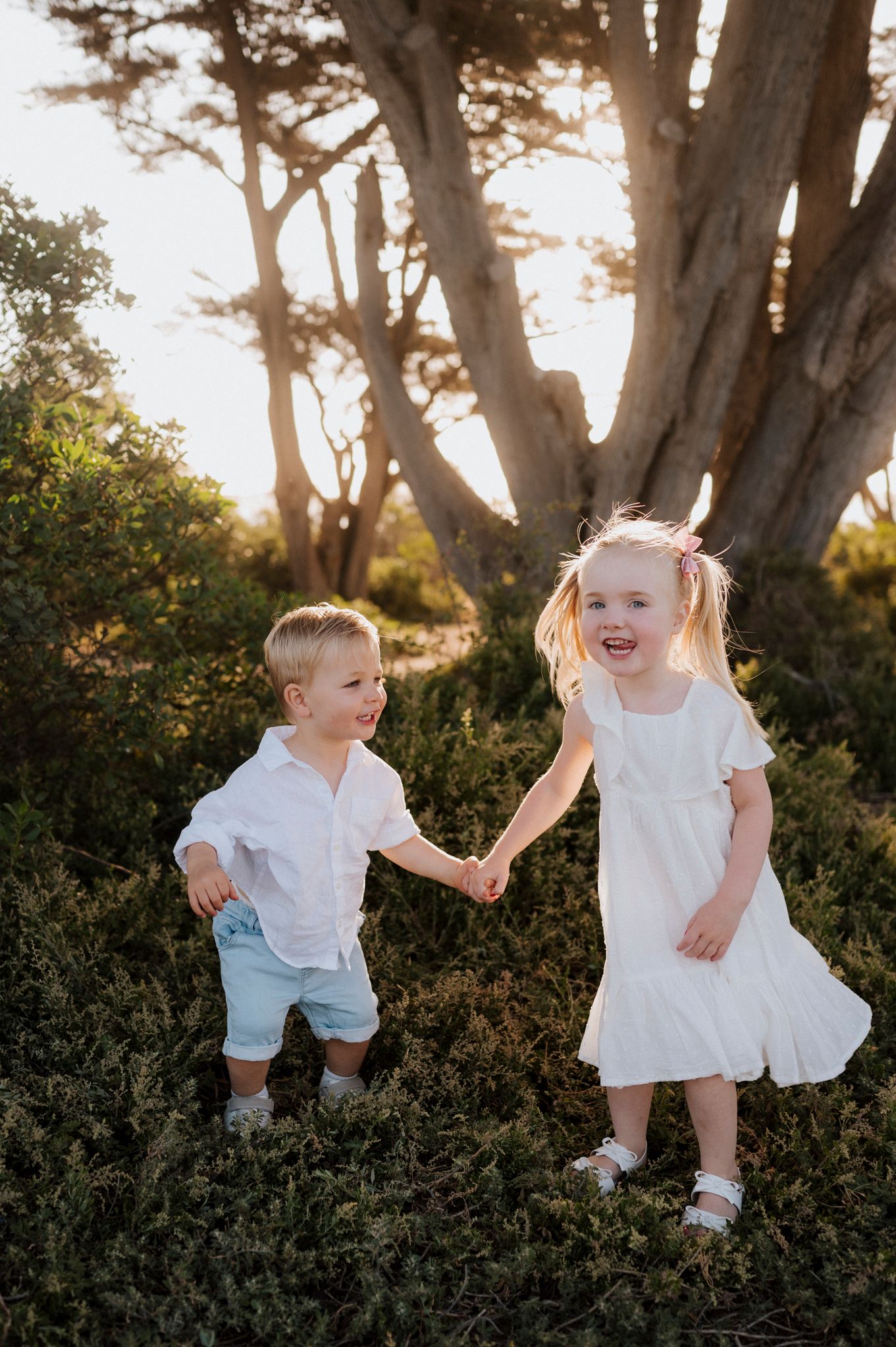 Two young children in white outfits hold hands and laugh while playing outdoors among green bushes and trees.