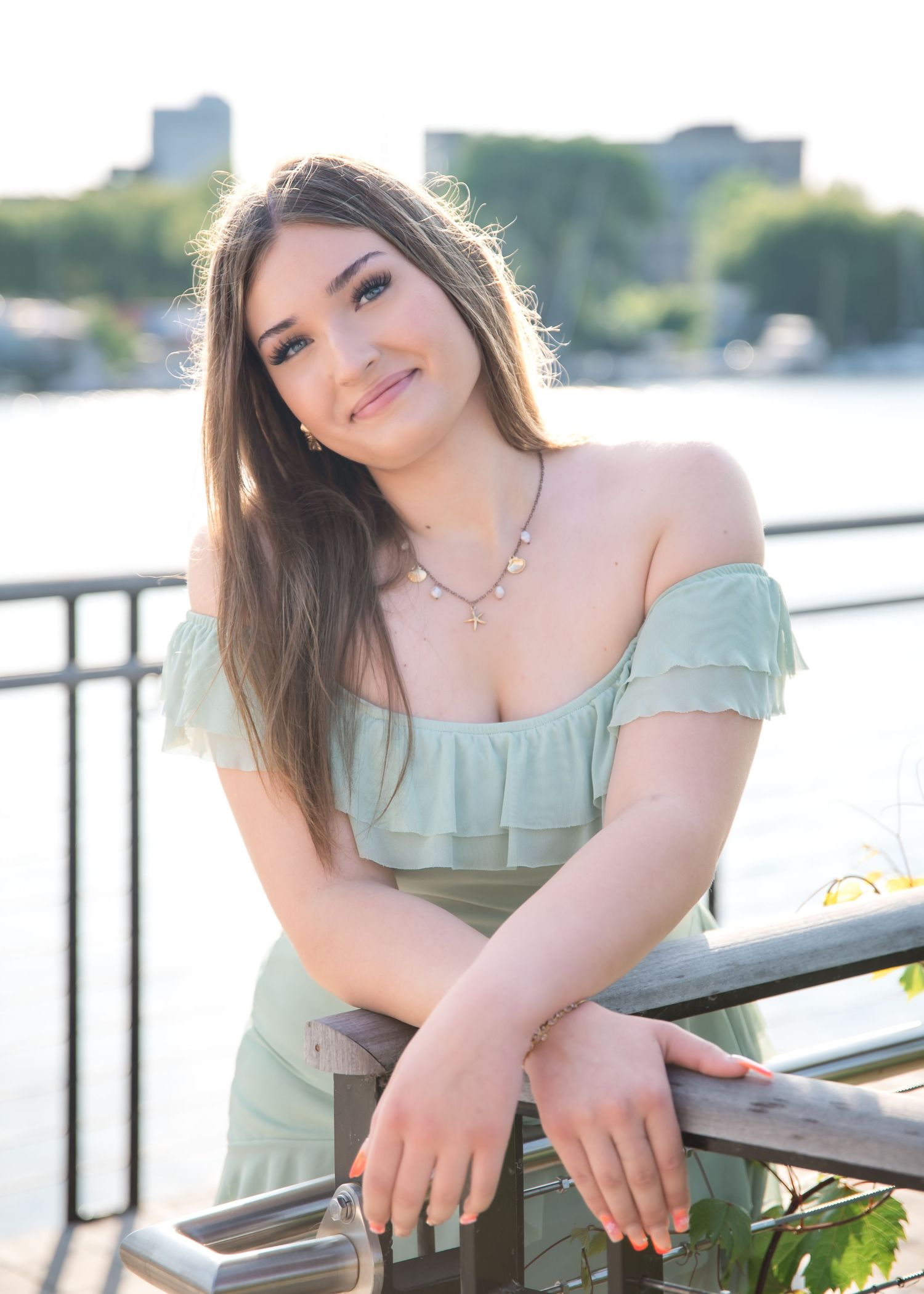 Young woman in sage green off-shoulder dress smiles on waterfront railing on a sunny day.