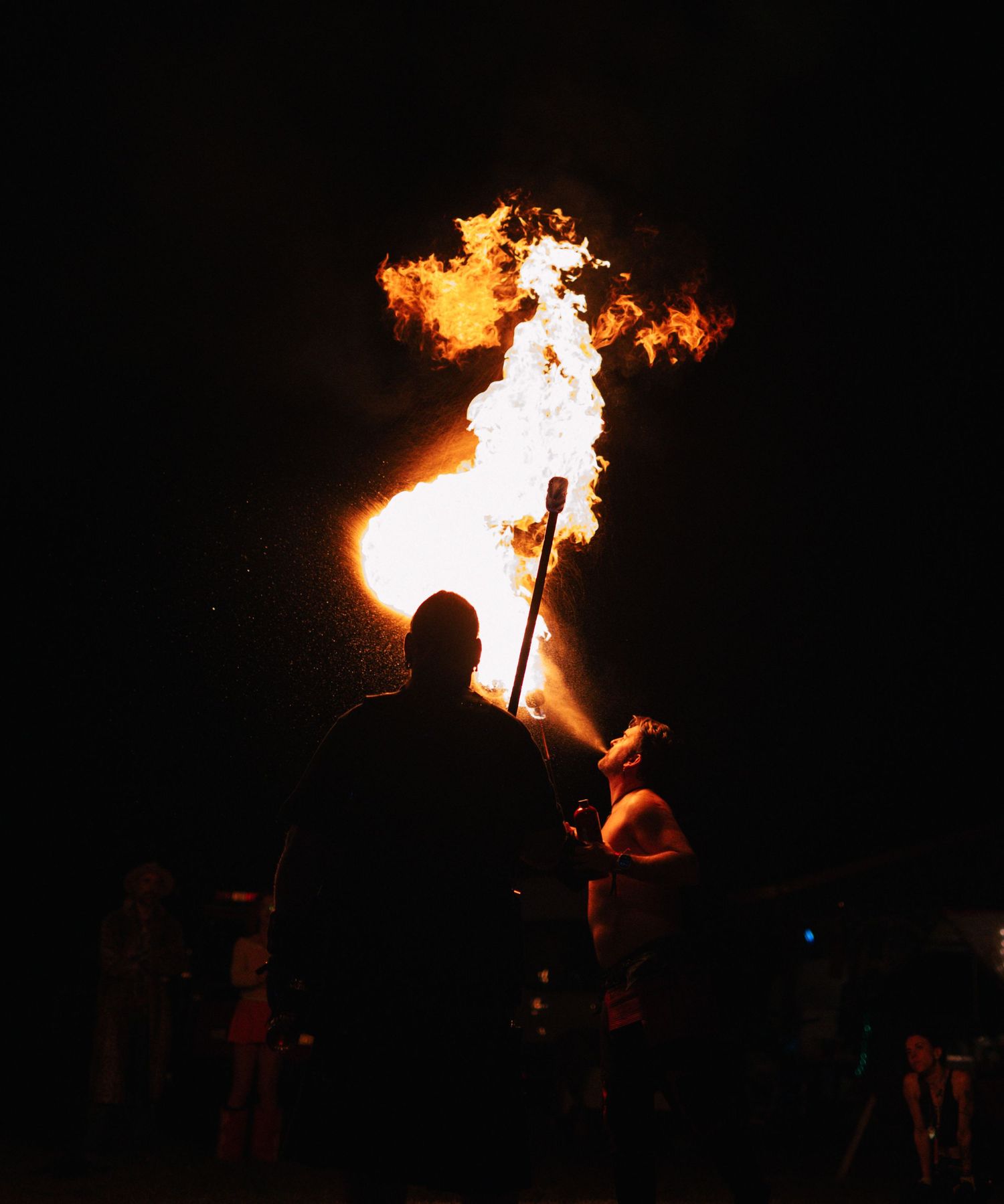 Ornate metal fire sculpture with tall flames at dusk, surrounded by seated spectators in a wooded outdoor setting.