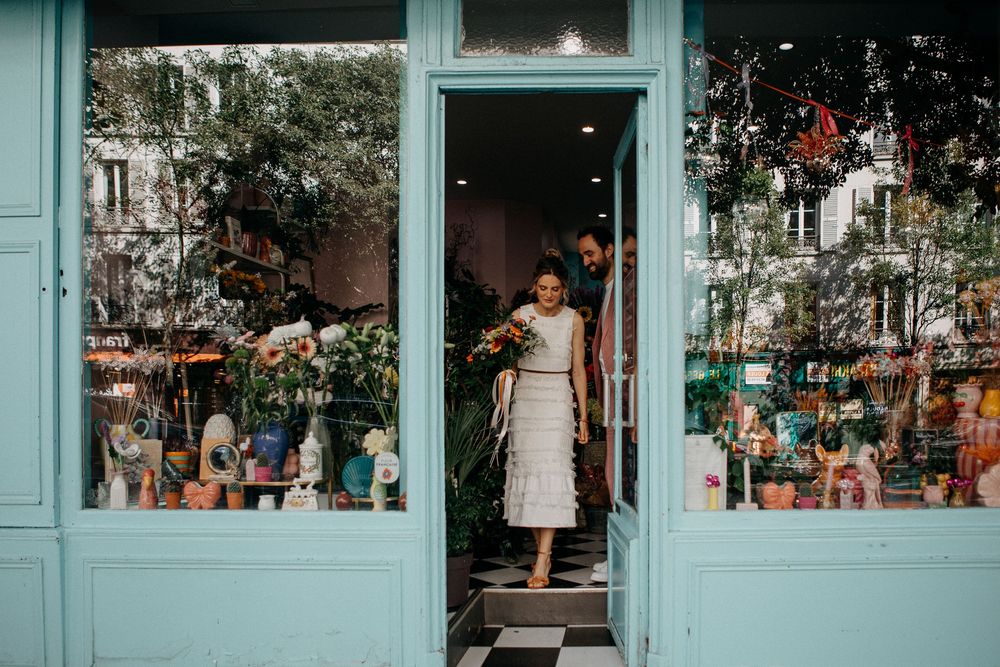 A bride in a white lace dress exits a charming turquoise flower shop doorway holding a colorful bouquet in Paris.
