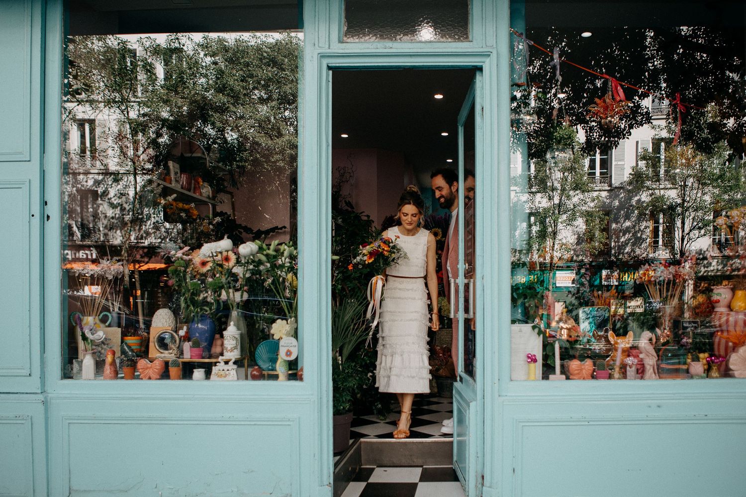 A bride in a white lace dress exits a charming turquoise flower shop doorway holding a colorful bouquet in Paris.