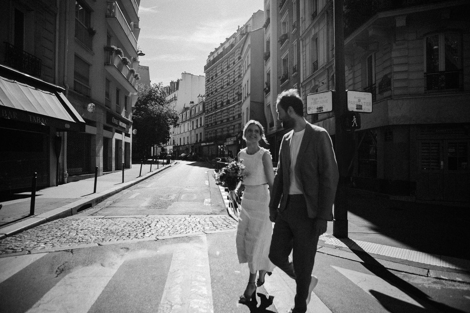 Black and white photo of a couple crossing a Parisian street, surrounded by classic Haussmann buildings.
