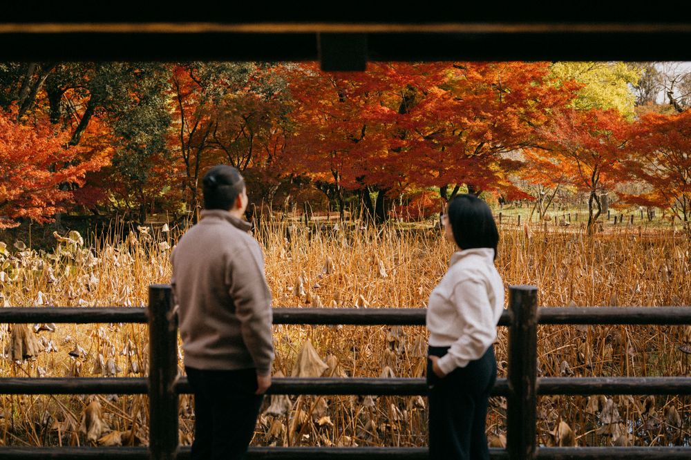 Couple standing side by side on wooden bridge viewing vibrant autumn foliage in golden orange hues.