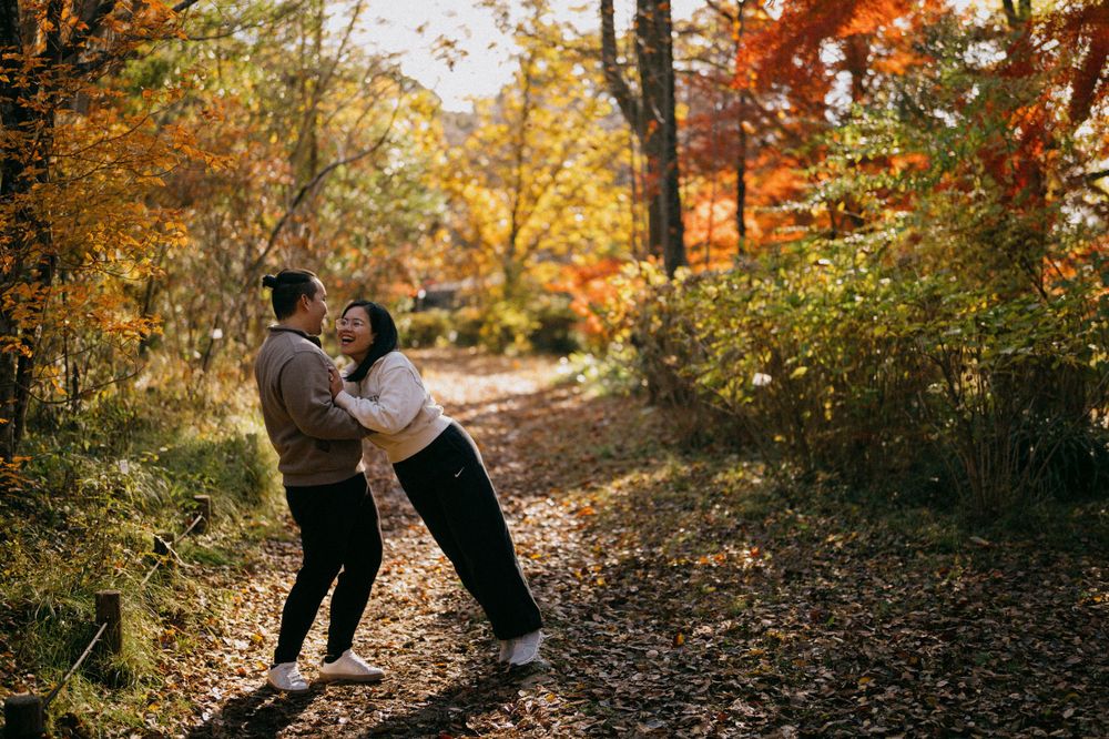 Couple playfully embracing and laughing together on a leaf-covered trail amid vibrant autumn forest.