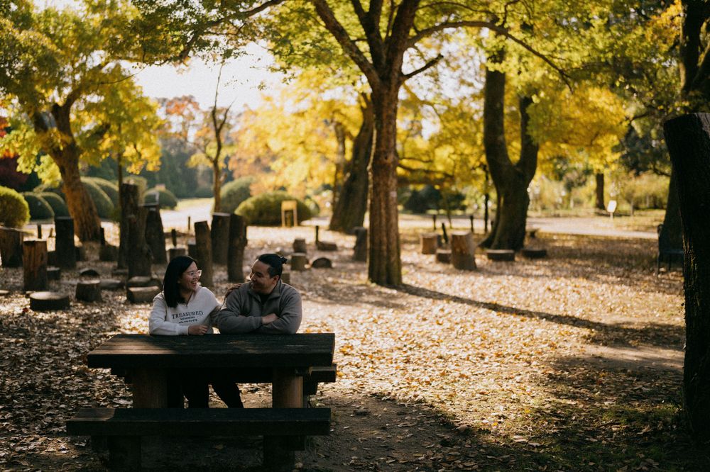 Couple sitting together at a picnic table in an autumn park surrounded by golden yellow foliage.
