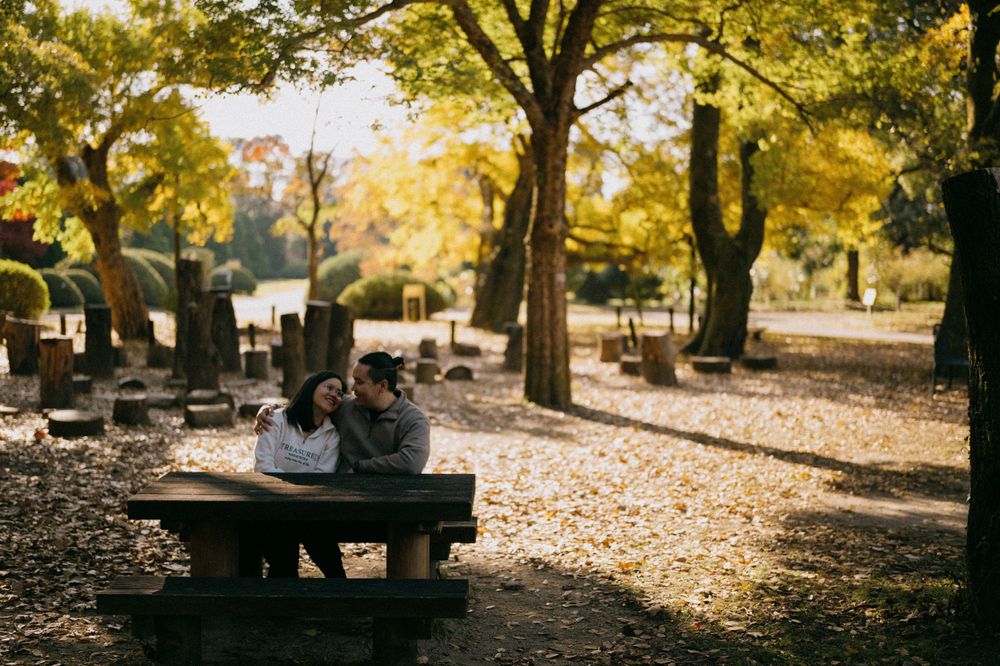 Two people share an intimate moment at a wooden bench amid fall leaves and golden trees.