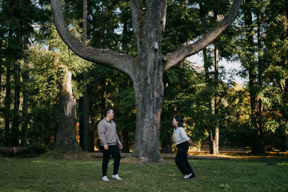 Couple playfully facing each other on a green lawn in front of a large branching tree.