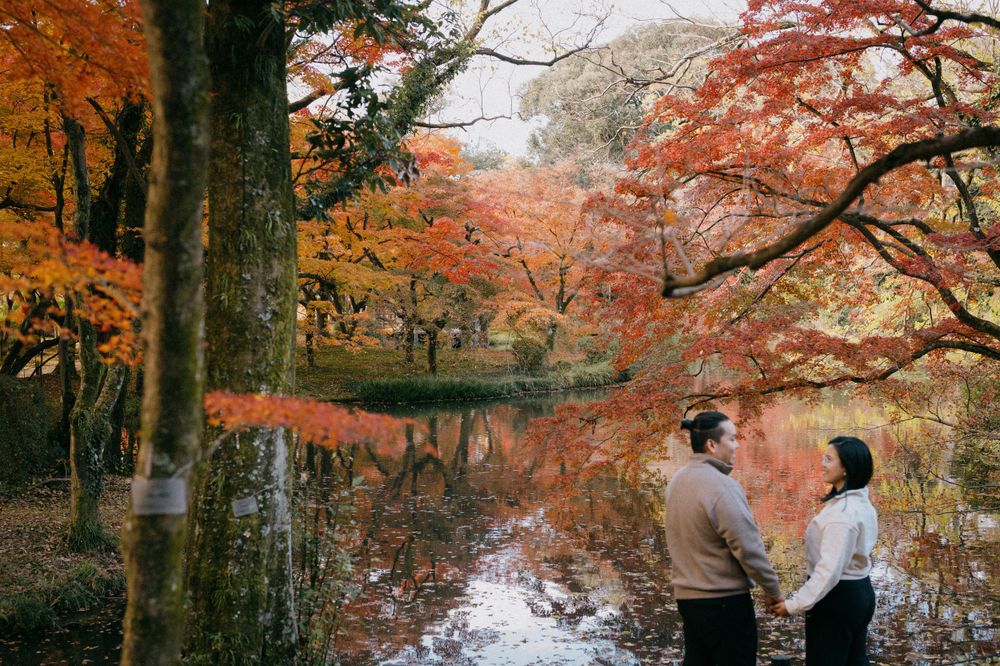 Couple standing hand in hand beside a tranquil pond with vivid autumn foliage reflections.