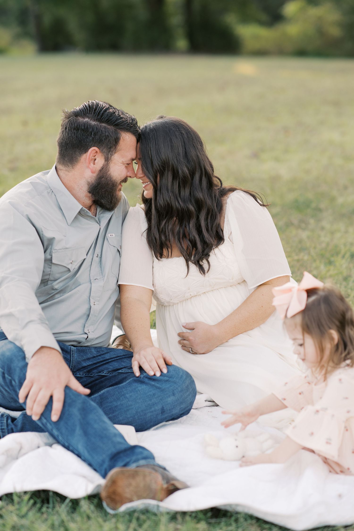 Pregnant couple posing in white studio, woman in mauve maternity gown, man in white shirt and khakis.