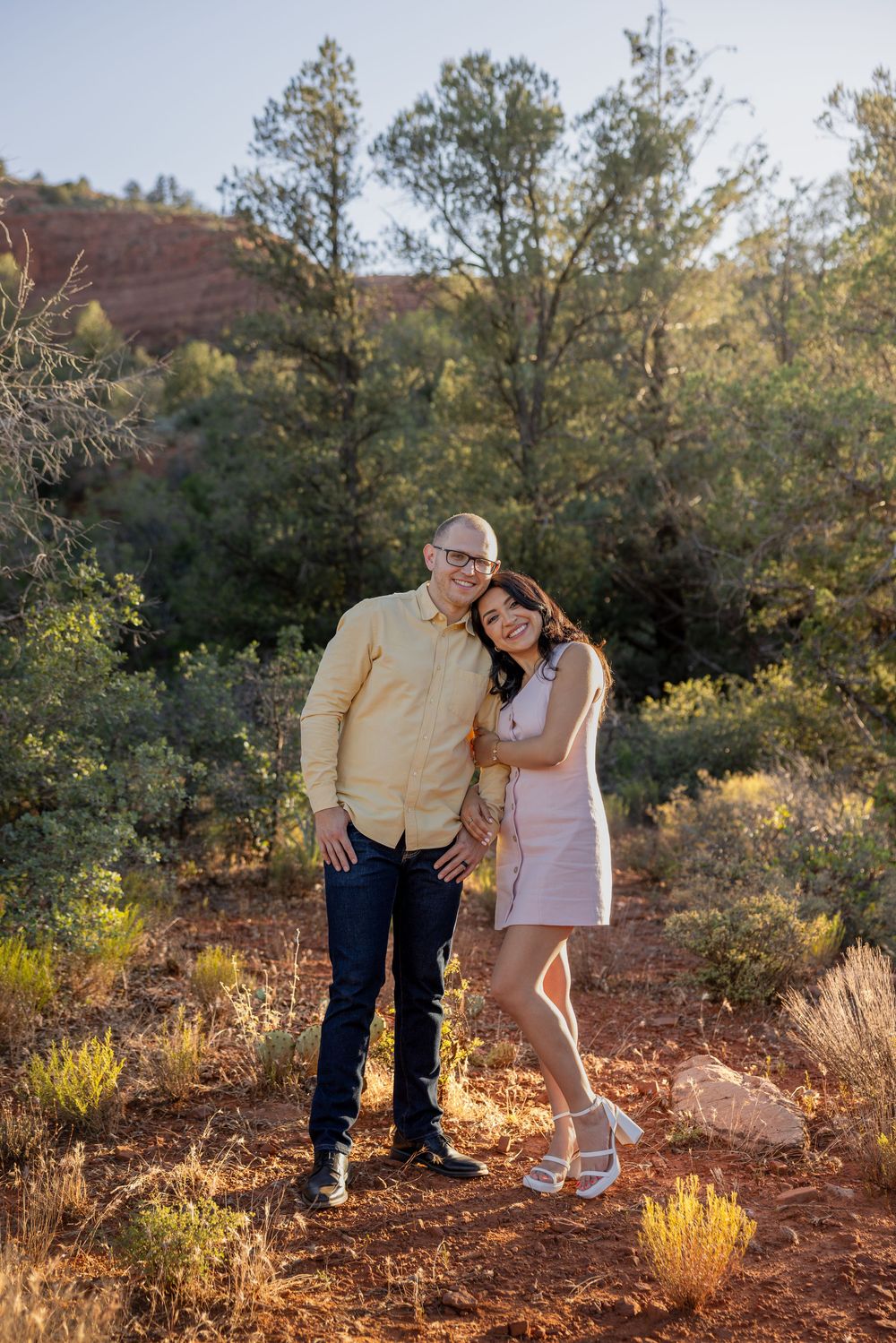 Couple embracing and smiling together among desert shrubs and red rock landscape in Sedona, Arizona.