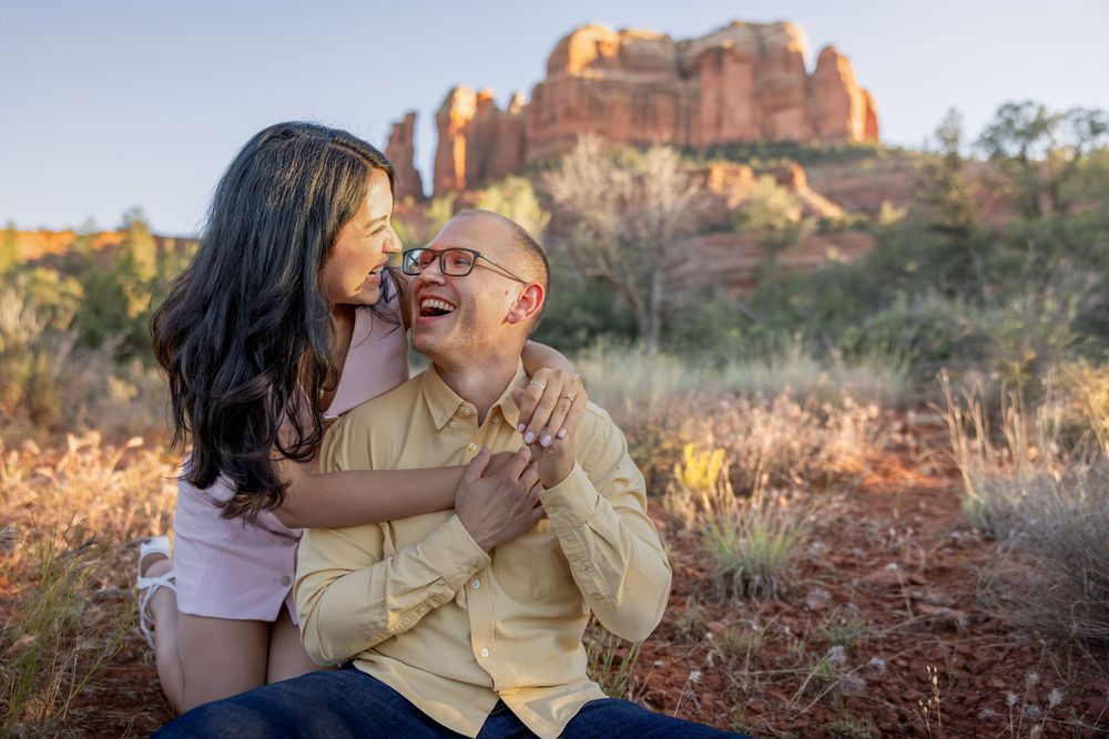 Couple laughing together in Sedona with red rock formations glowing at sunset behind them.