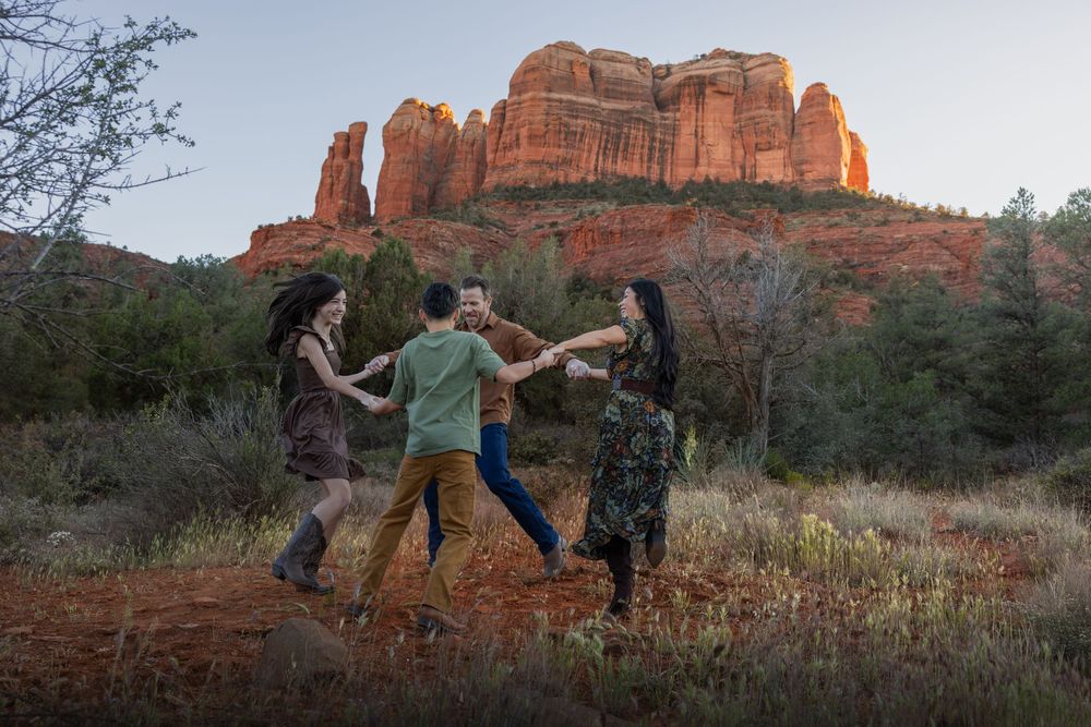 Family of four joyfully dancing in a circle on red soil with Cathedral Rock at sunset.