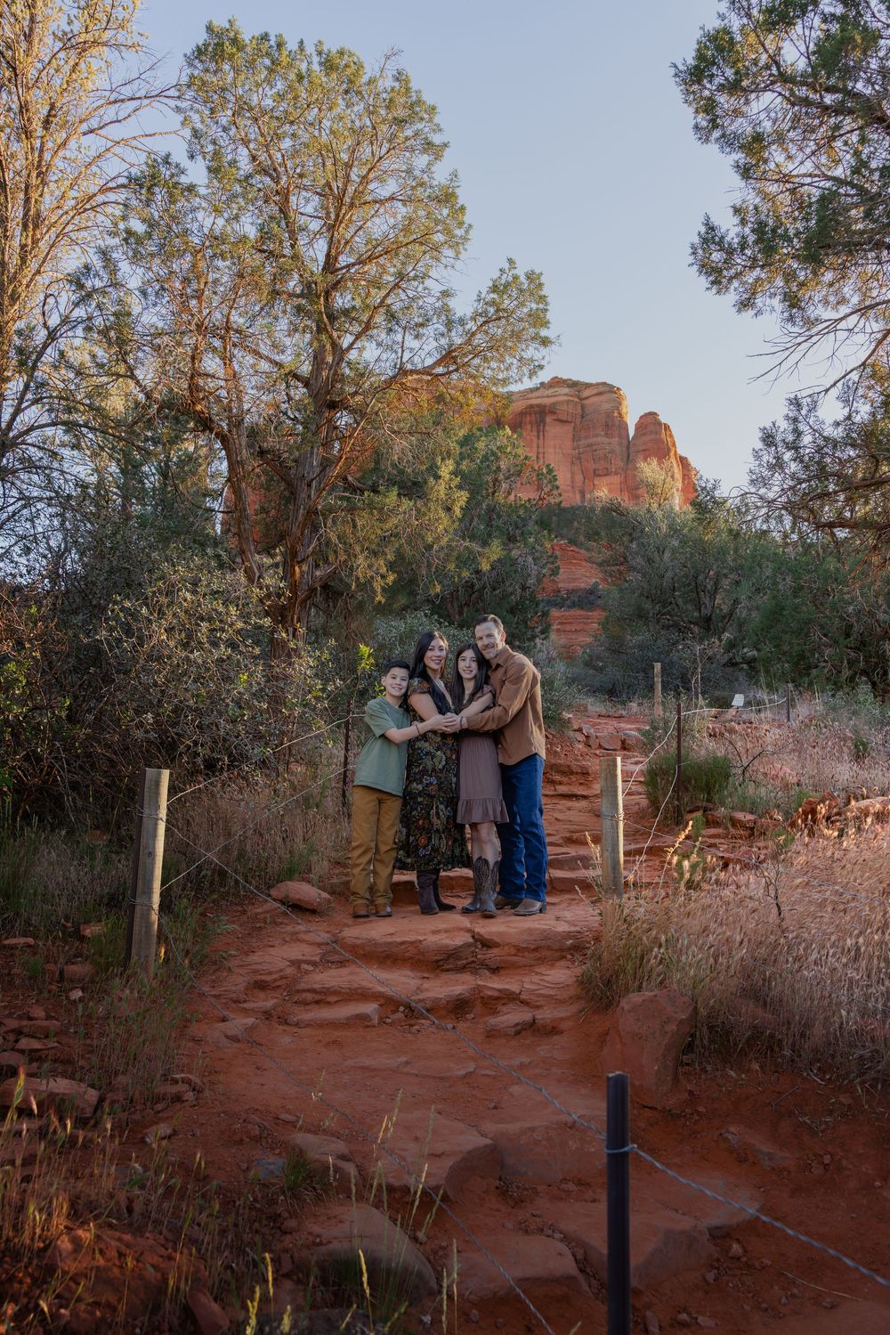 Family of four hugging on a red dirt trail with Cathedral Rock in Sedona at dusk.