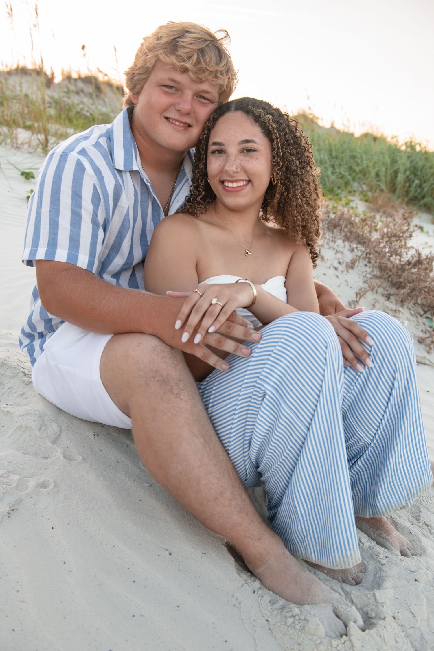 A young couple sits together on sandy beach dunes, smiling warmly, dressed in coordinating blue and white outfits.