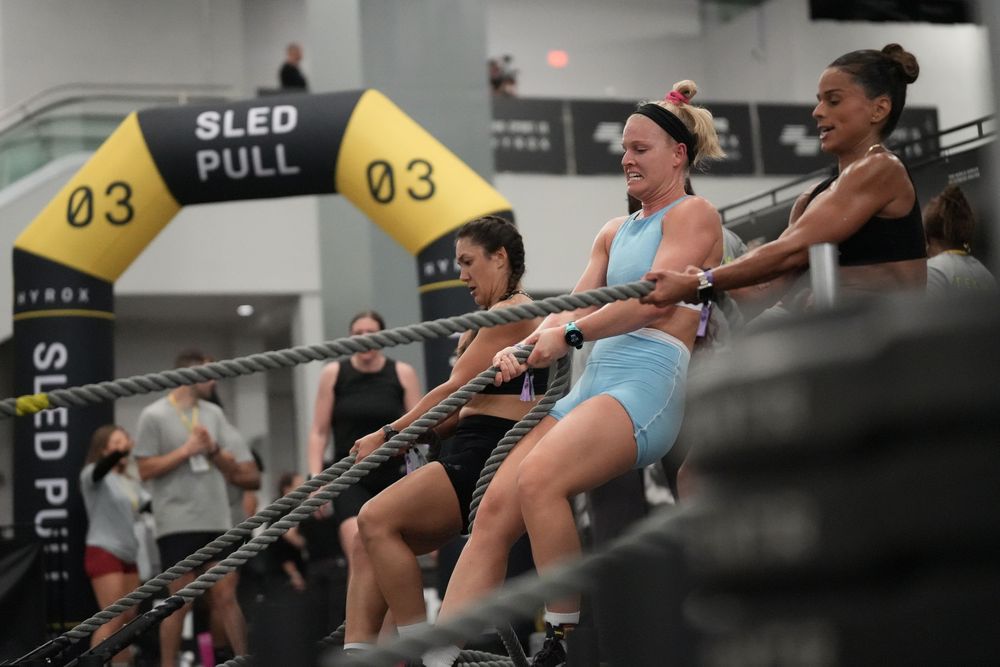 Hyrox athletes competing at the sled pull station, showcasing strength, teamwork, endurance, and functional fitness race action.