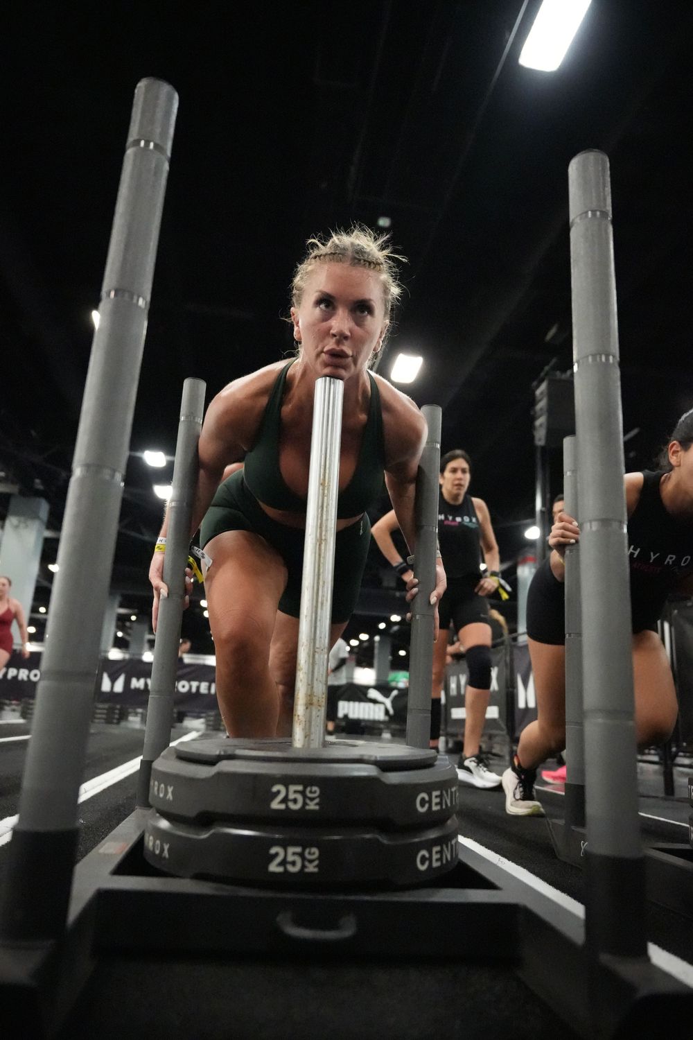 Low-angle action photograph of a female athlete powering through the Hyrox sled push station, emphasizing strength, determination