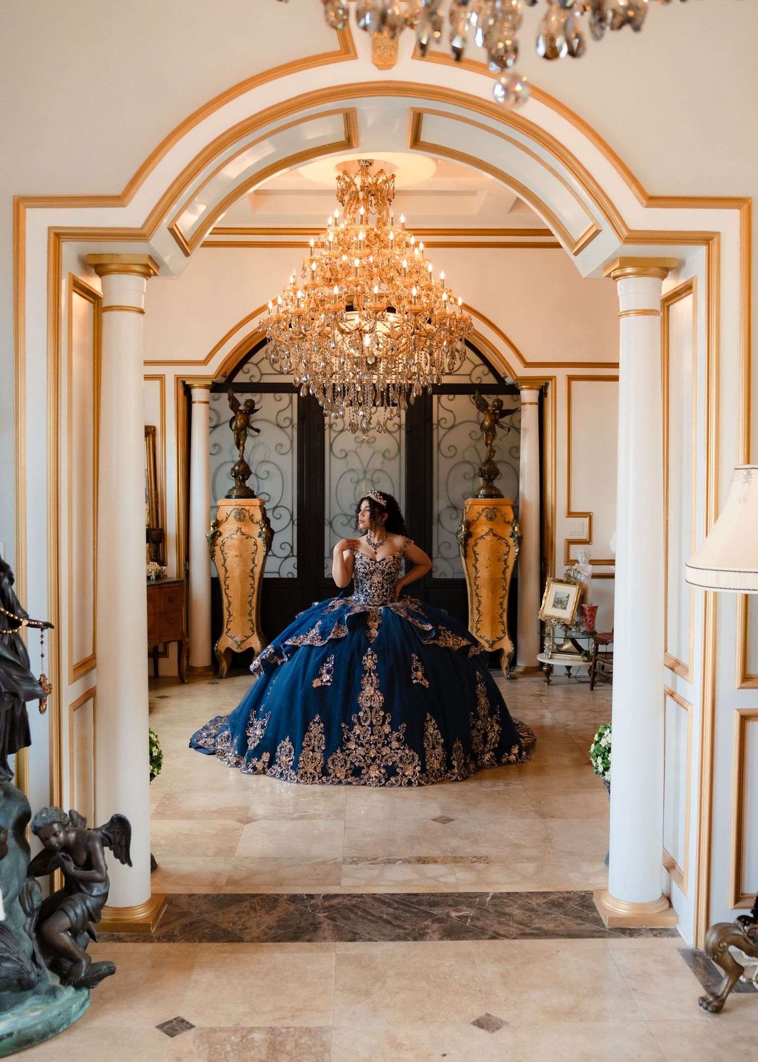 Young woman in navy blue and gold quinceañera ball gown stands under crystal chandelier in elegant mansion foyer.