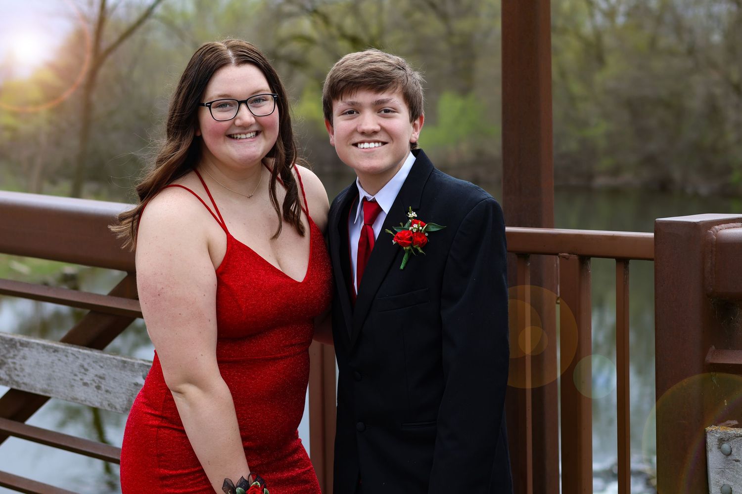 A smiling couple dressed in formal prom attire, red dress and black suit, posing on a wooden bridge outdoors.