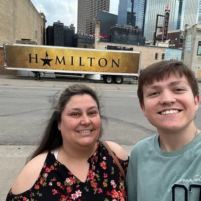 Two people smiling in front of a Hamilton musical tour truck with city skyline in background.