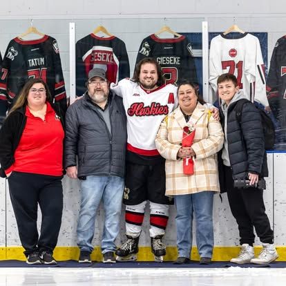 A family of five poses together on ice rink with hockey jerseys displayed on the boards behind them.