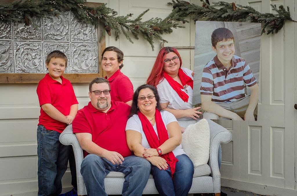 Family of five poses indoors with holiday greenery, wearing red and white, with a framed portrait nearby.