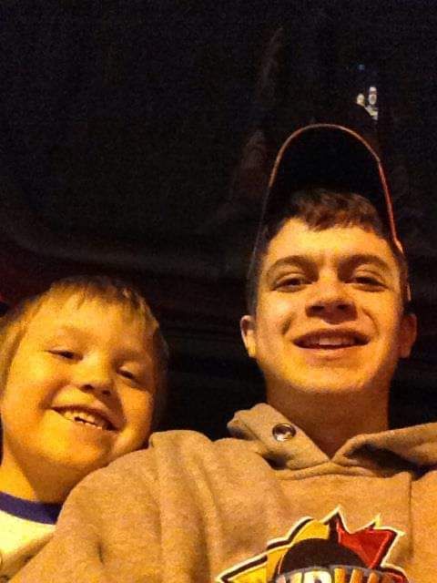 Two smiling boys taking a selfie together, the younger boy on the left and older teen wearing a hoodie on the right.