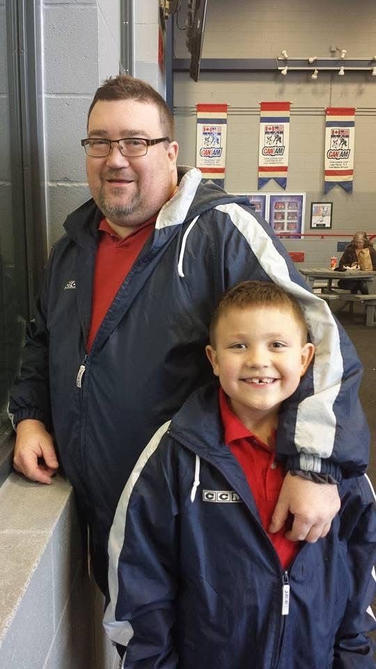 A man with glasses and a young boy both wearing navy blue jackets smile together inside a hockey arena.