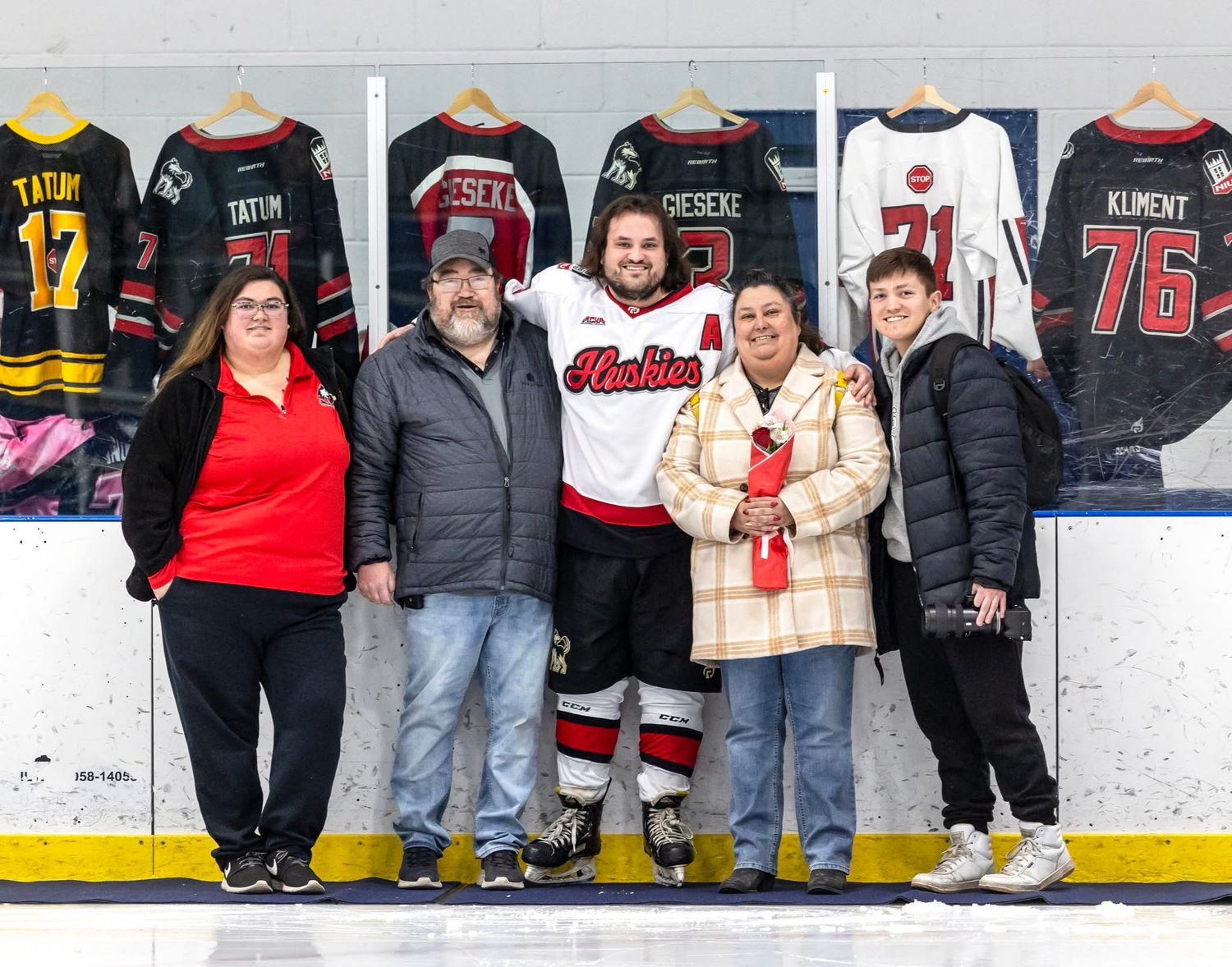Hockey player in uniform poses with four family members on ice rink, jerseys displayed on boards behind them.