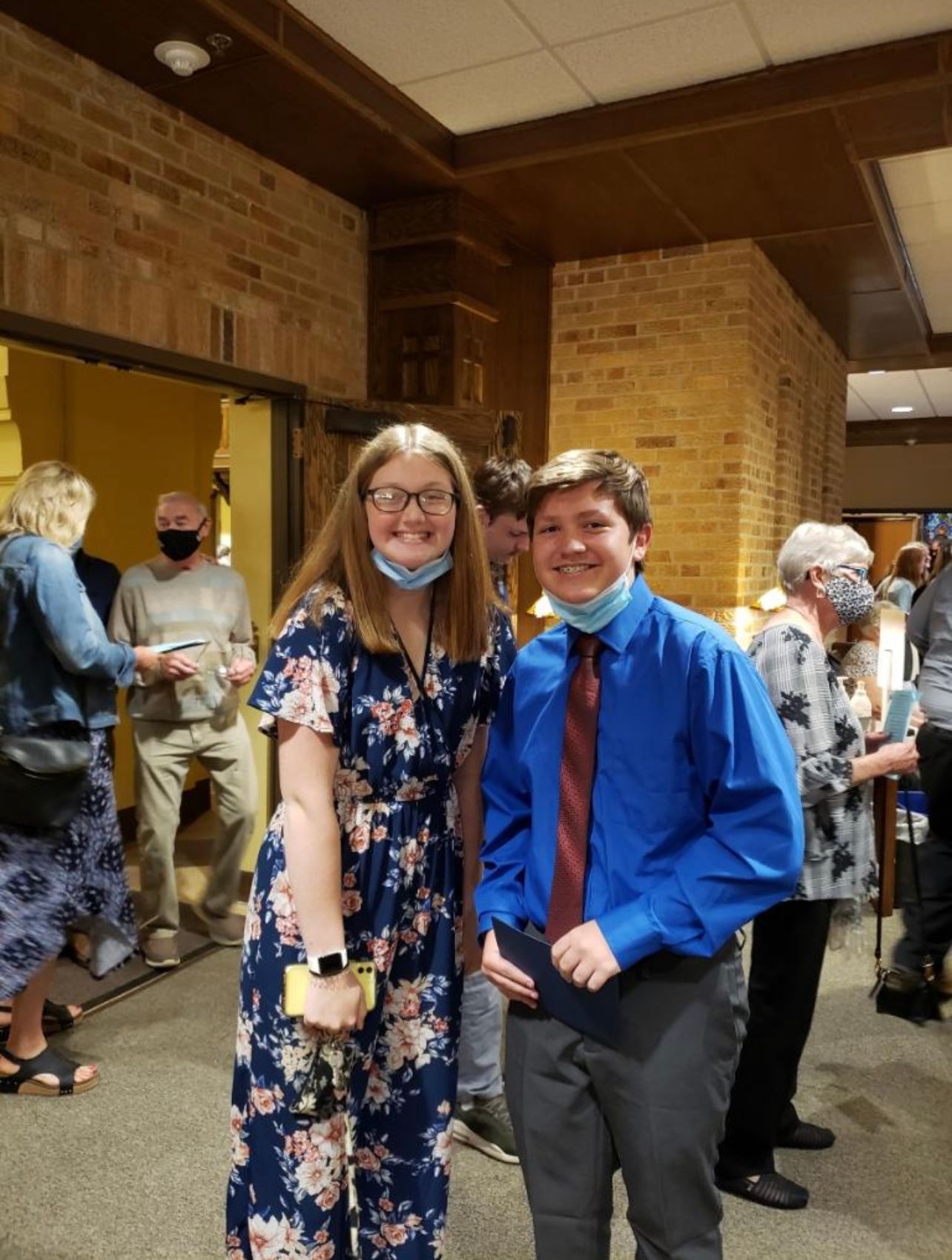 Two young people in formal attire wearing masks pose together at an indoor event or ceremony.