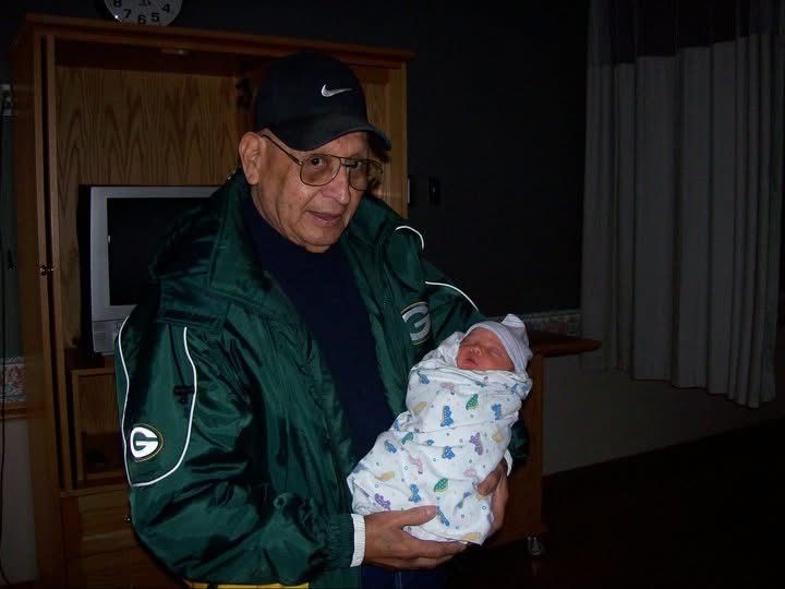 Elderly man in green jacket holds a sleeping newborn baby wrapped in a white blanket indoors.