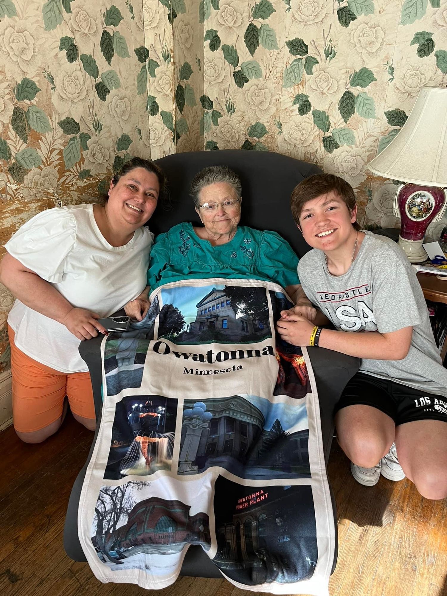 Two women and an elderly lady display an Owatonna, Minnesota souvenir blanket while sitting together indoors.
