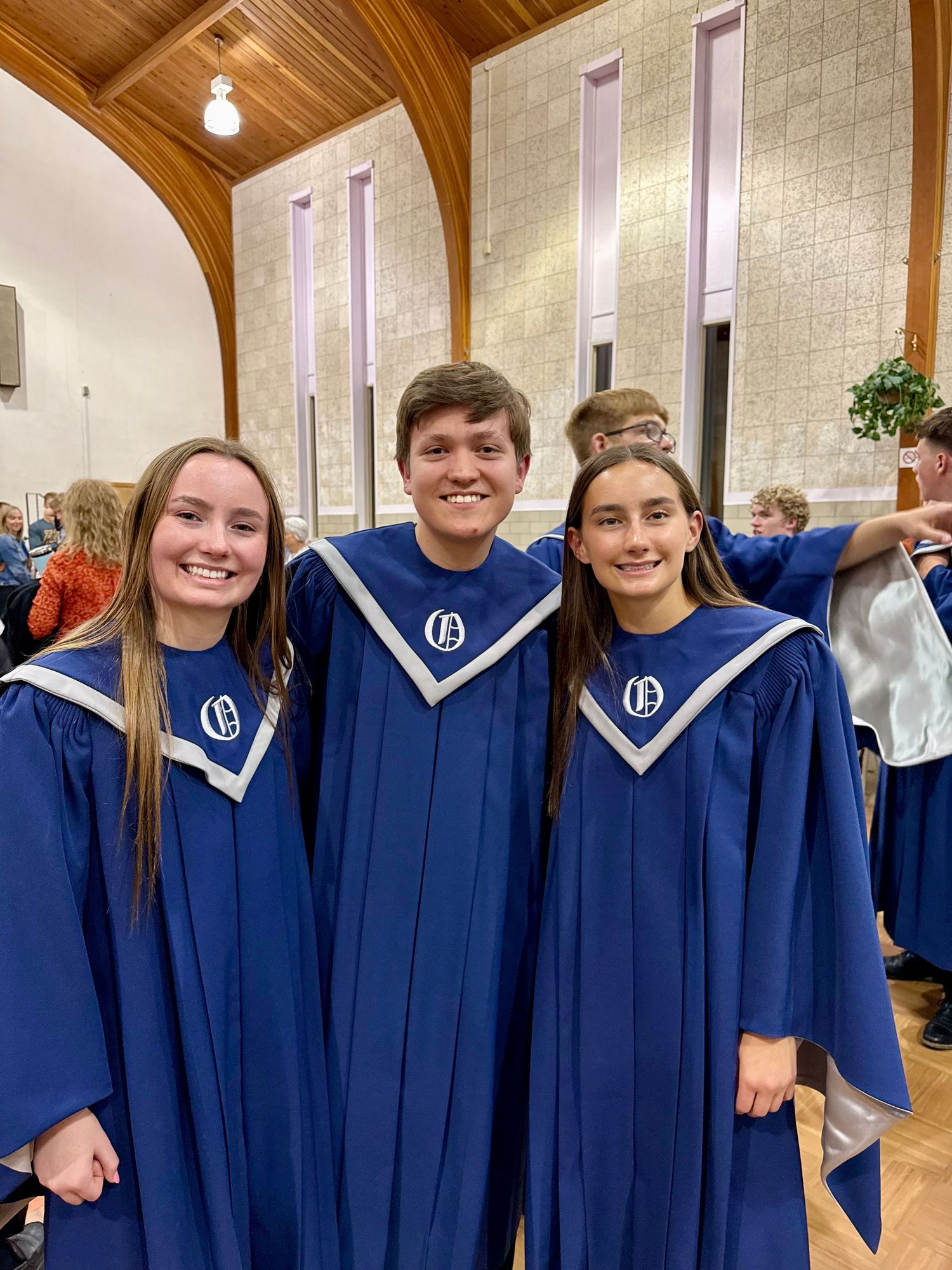 Three smiling students wearing blue choir robes with emblem stand together in a church during a choral performance.