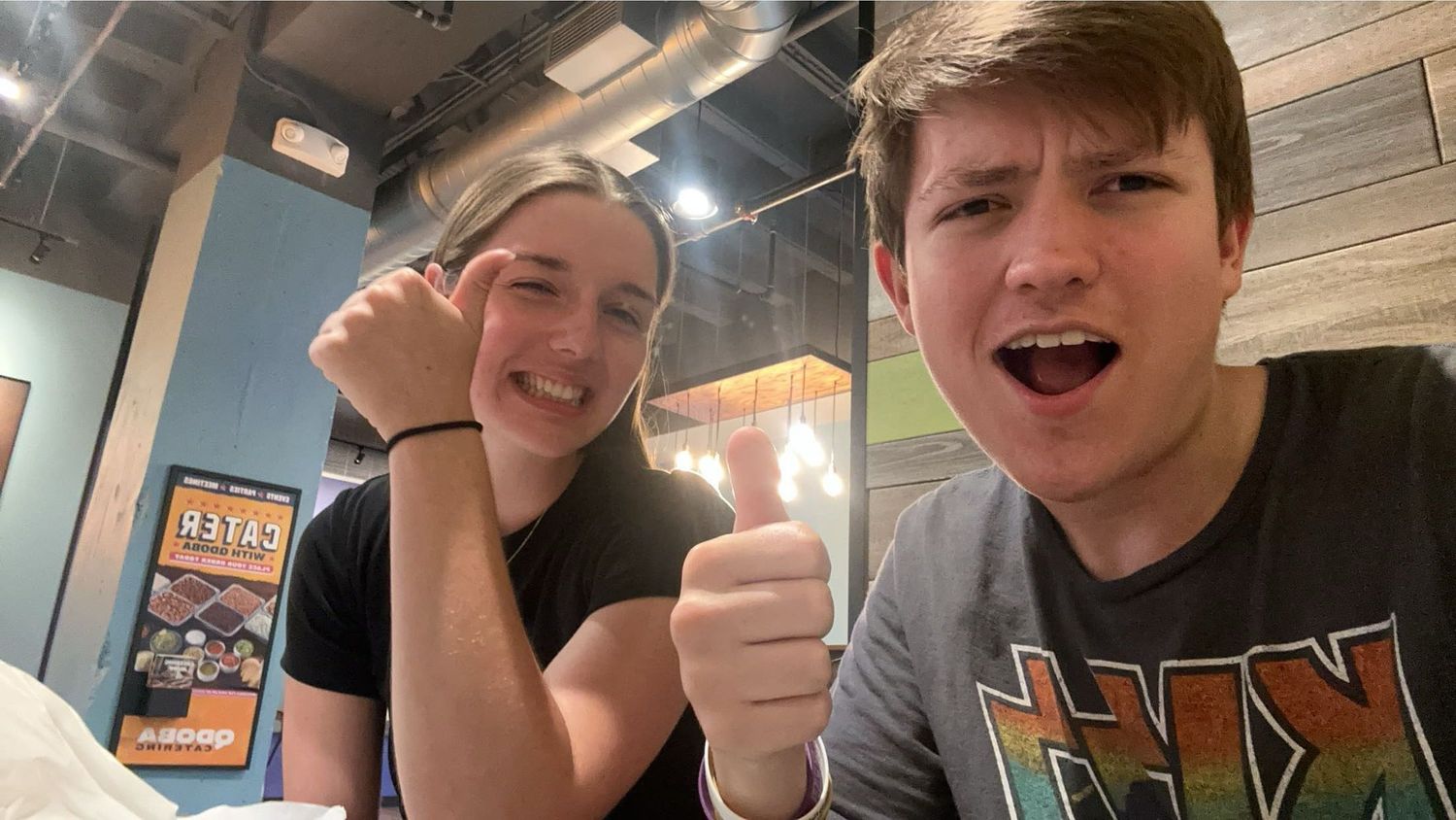 Two smiling young adults giving thumbs up at a casual restaurant, posing happily for a selfie photo.